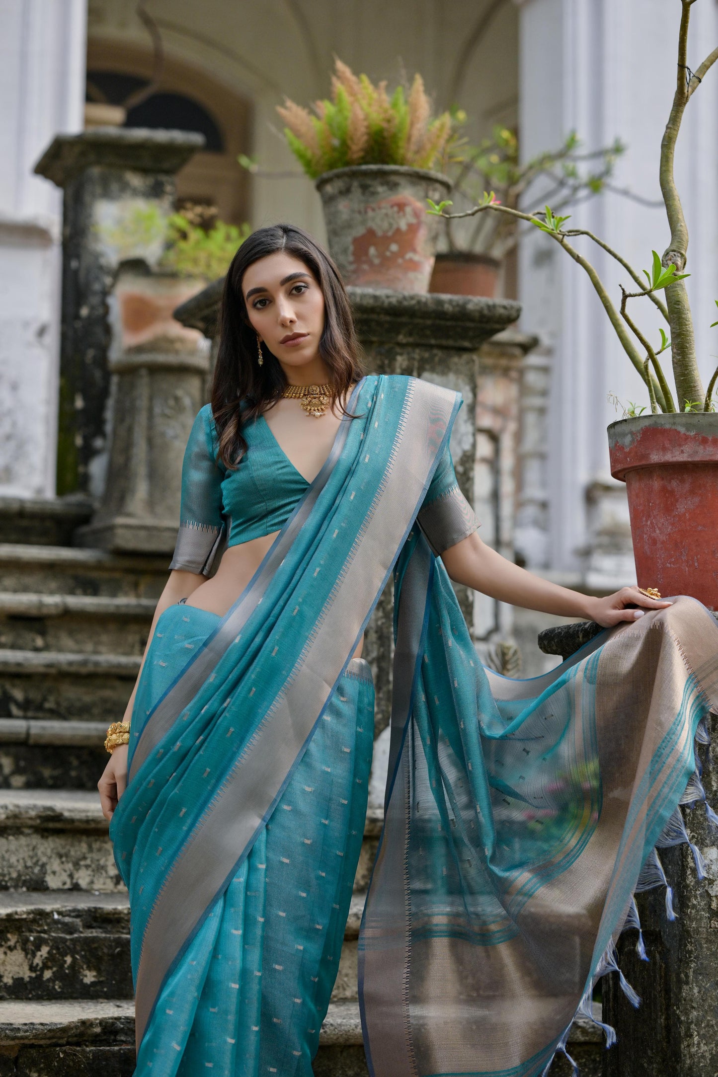 Woman wearing a teal silk saree with a silver border and subtle woven motifs, paired with a matching blouse and gold jewelry, standing on the steps of a heritage mansion surrounded by potted plants and lush greenery.