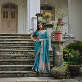 Woman wearing a teal silk saree with a silver border and subtle woven motifs, paired with a matching blouse and gold jewelry, standing on the steps of a heritage mansion surrounded by potted plants and lush greenery.