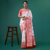 Woman wearing a white silk saree with coral pink and gold woven border, featuring circular and geometric motifs, paired with a matching coral blouse with white polka dots, accessorized with a choker necklace and green bangles, posing in a studio setting next to a wooden stool.