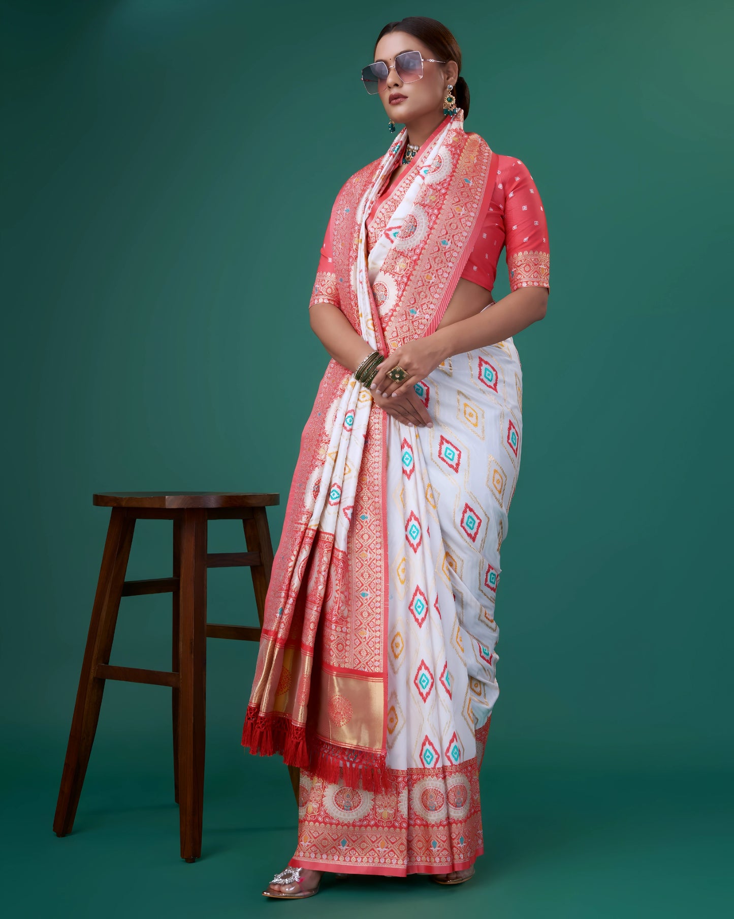 Woman wearing a white silk saree with coral pink and gold woven border, featuring circular and geometric motifs, paired with a matching coral blouse with white polka dots, accessorized with a choker necklace and green bangles, posing in a studio setting next to a wooden stool.