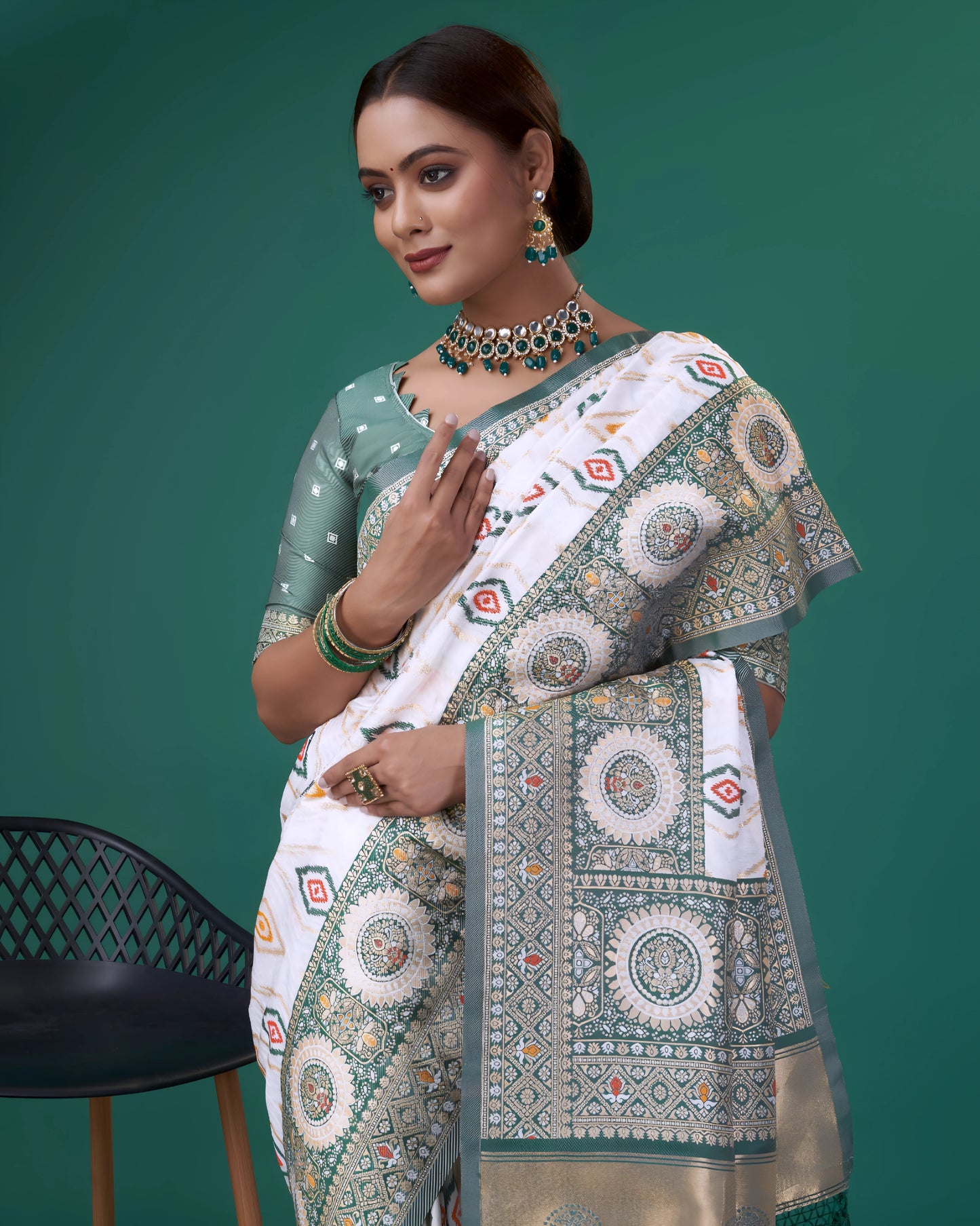 Woman wearing a white silk saree with green and gold woven border, featuring circular and diamond-shaped geometric motifs, paired with a green polka dot blouse and traditional jewelry, posing in a studio setting next to a modern black chair.