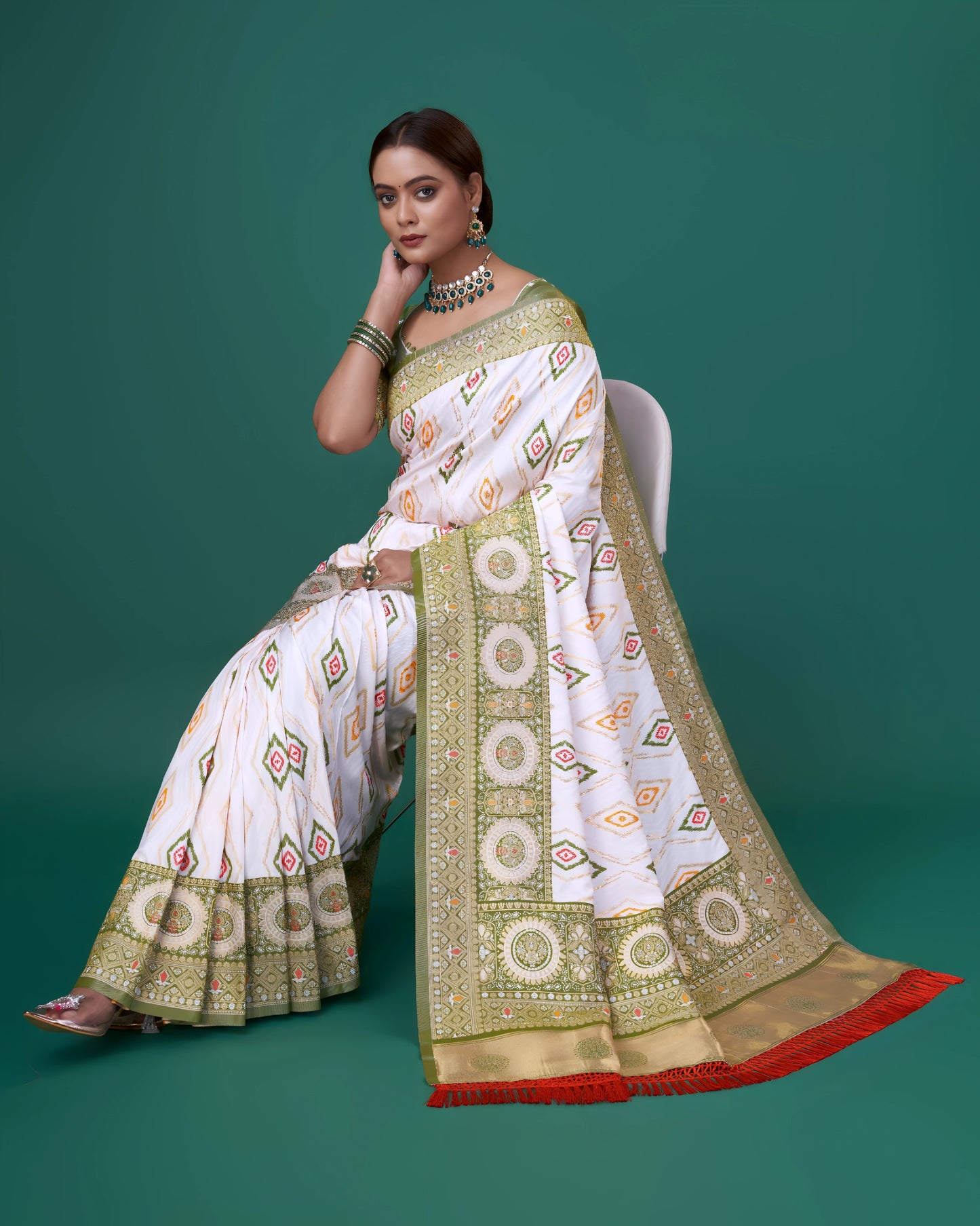 Woman wearing a white silk saree with intricate green and gold border, featuring circular and diamond-shaped geometric motifs, paired with a green polka dot blouse and traditional jewelry, standing beside a wooden stool in a studio with a green backdrop.