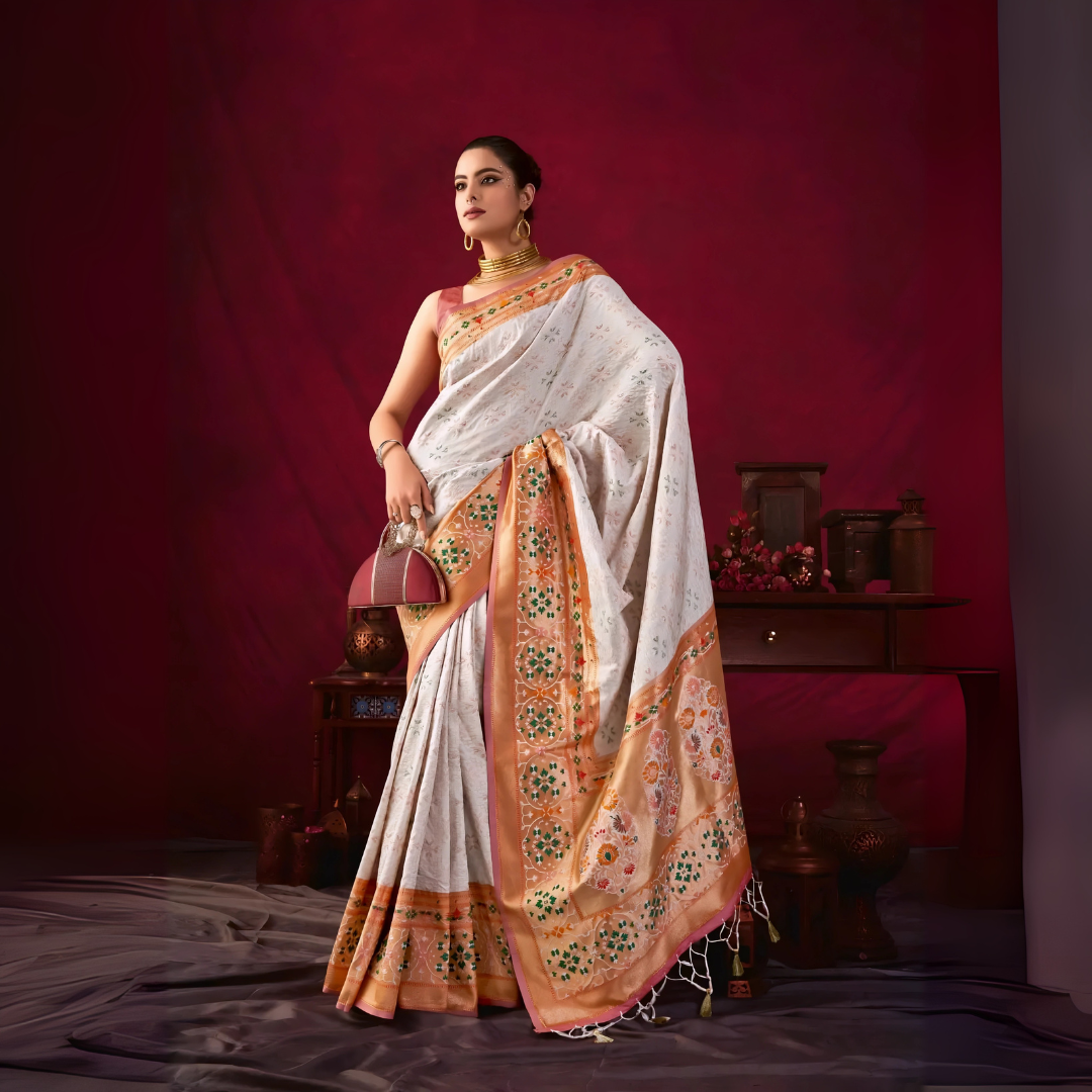 Woman wearing a white silk saree with an orange and gold floral embroidered border, accessorized with gold jewelry and a maroon handbag, standing in front of a vintage wooden table and decor against a deep red backdrop.