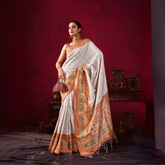 Woman wearing a white silk saree with an orange and gold floral embroidered border, accessorized with gold jewelry and a maroon handbag, standing in front of a vintage wooden table and decor against a deep red backdrop.