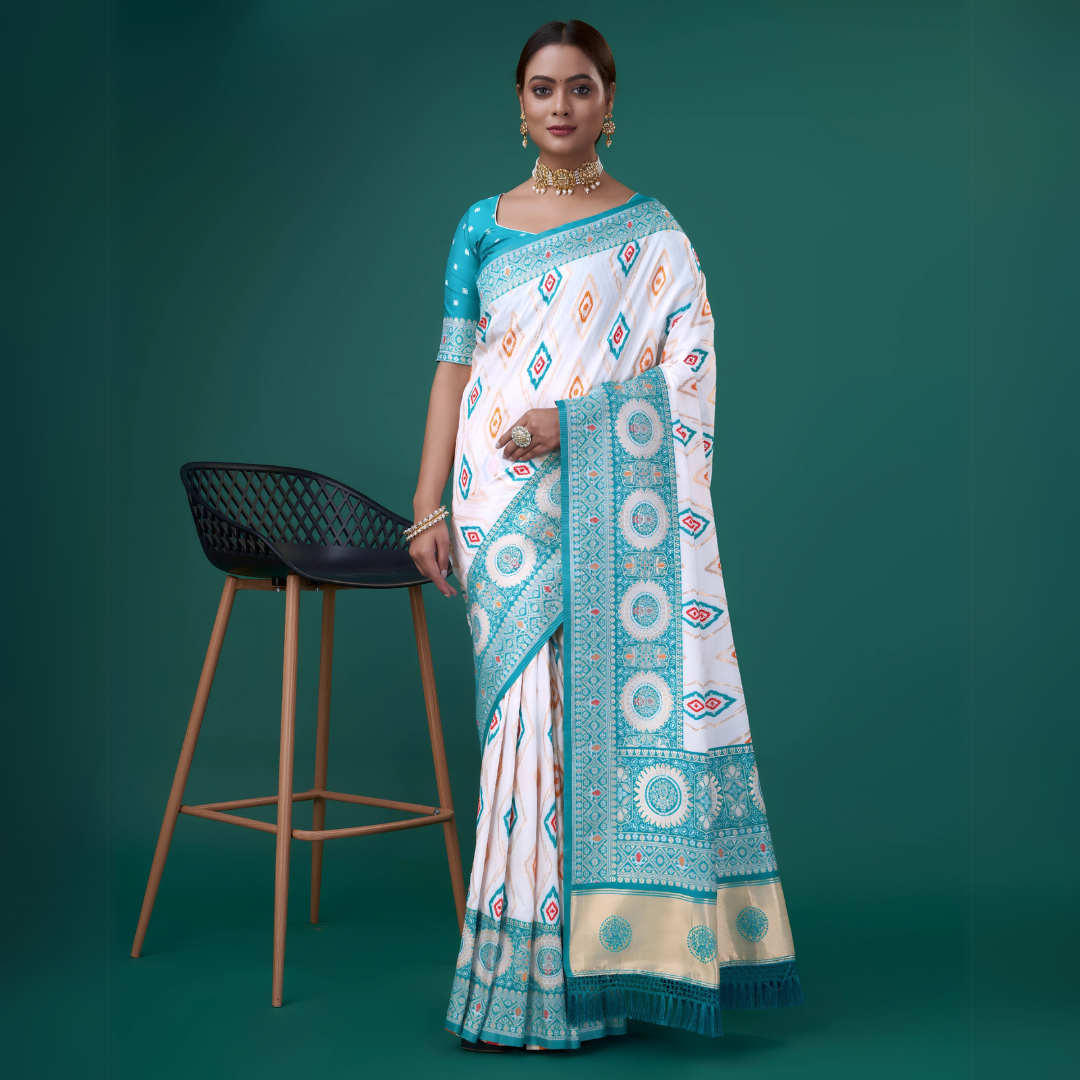 Woman wearing a white silk saree with turquoise blue border and intricate circular and diamond-shaped geometric motifs, paired with a matching turquoise blouse adorned with polka dots, accessorized with traditional jewelry, posing beside a modern black chair in a studio with a green backdrop.
