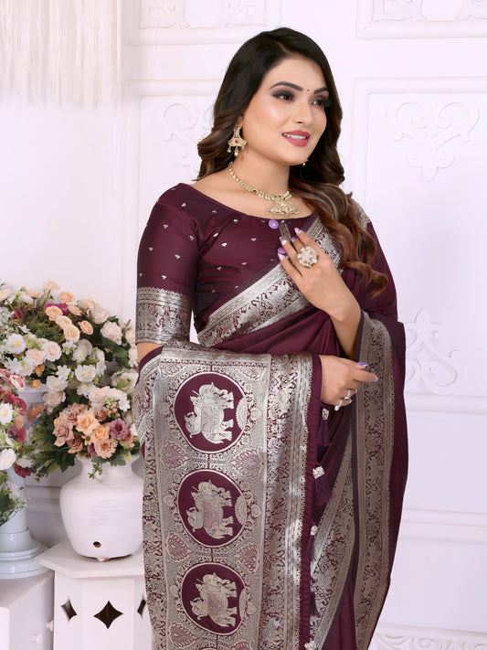 Woman wearing a wine-colored silk saree adorned with an elegant silver zari border and traditional elephant motifs on the pallu, paired with a matching blouse featuring silver detailing. She poses gracefully in a classic white floral-themed room decorated with ornate wall panels and flower arrangements in white vases.