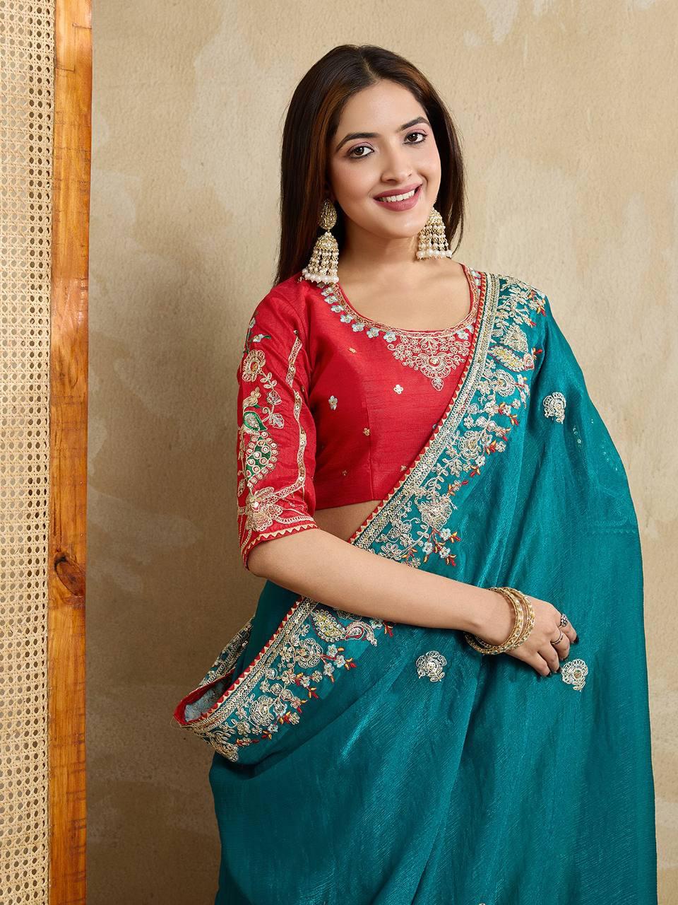 Woman wearing a teal blue embroidered saree with intricate floral border and motifs, paired with a red blouse and traditional jewelry, standing indoors beside a wooden chair and cane screen