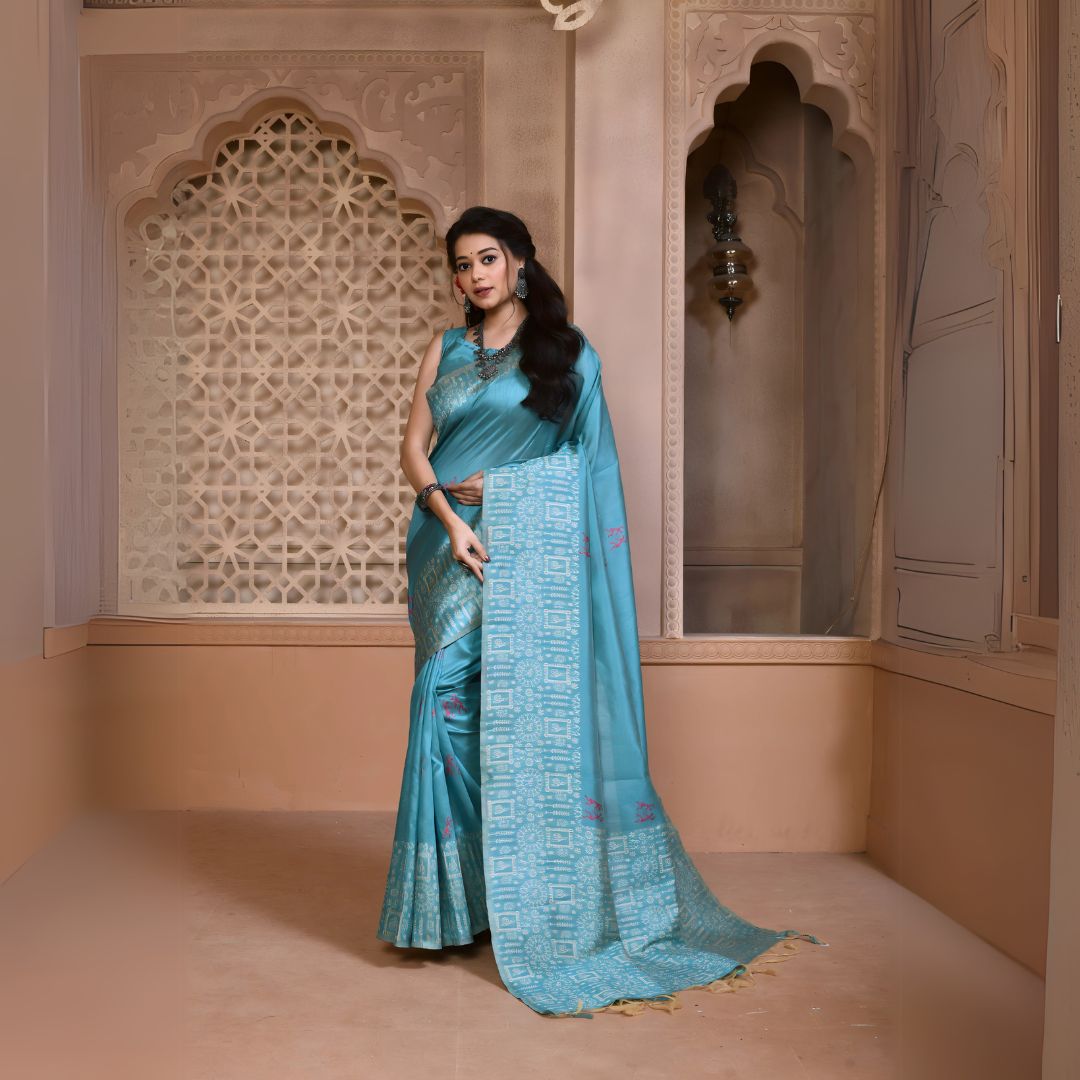 Woman wearing an aqua blue silk saree with geometric patterned border and pallu, accessorized with traditional jewelry, standing in a heritage-inspired room with ornate carved windows and pastel walls.