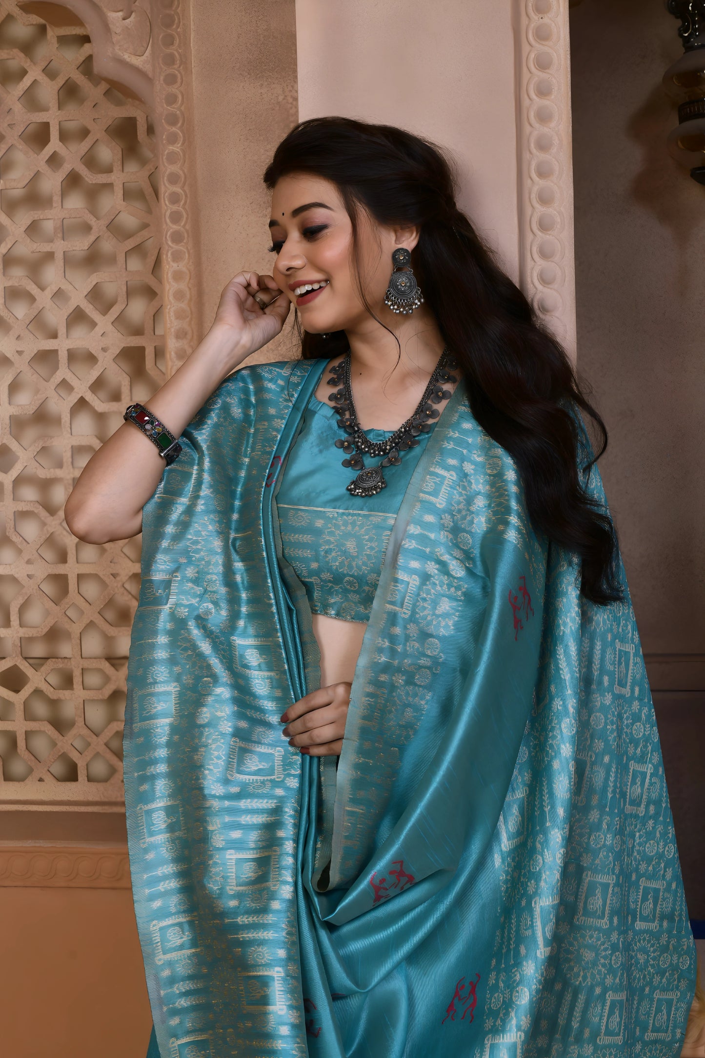 Woman wearing an aqua blue silk saree with geometric patterned border and pallu, accessorized with traditional jewelry, standing in a heritage-inspired room with ornate carved windows and pastel walls.