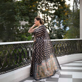 Woman wearing a black saree with bold beige geometric stripes and a floral printed pallu, standing on a balcony with decorative railing overlooking a lush green garden.