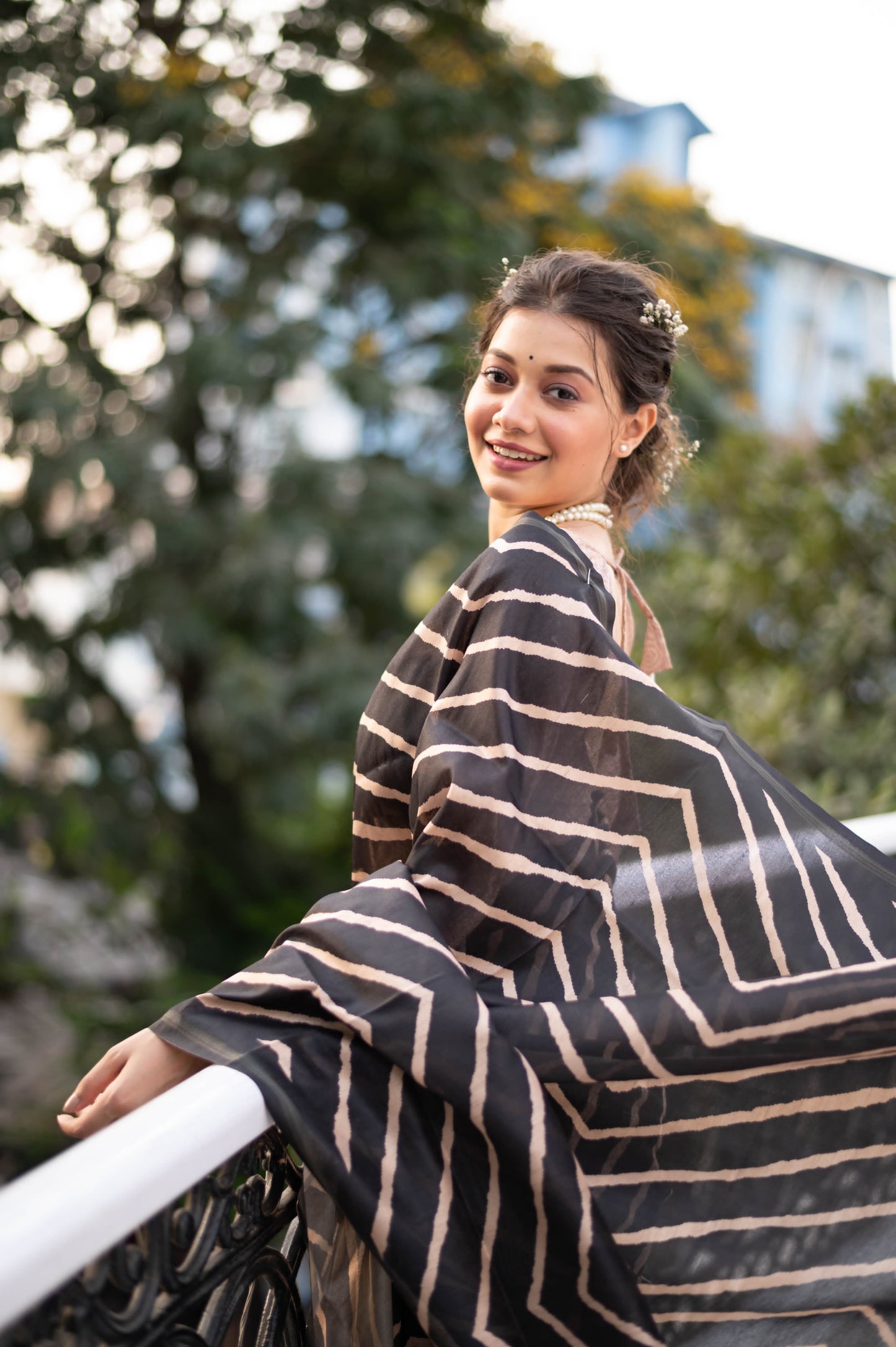Woman wearing a black saree with bold beige geometric stripes and a floral printed pallu, standing on a balcony with decorative railing overlooking a lush green garden