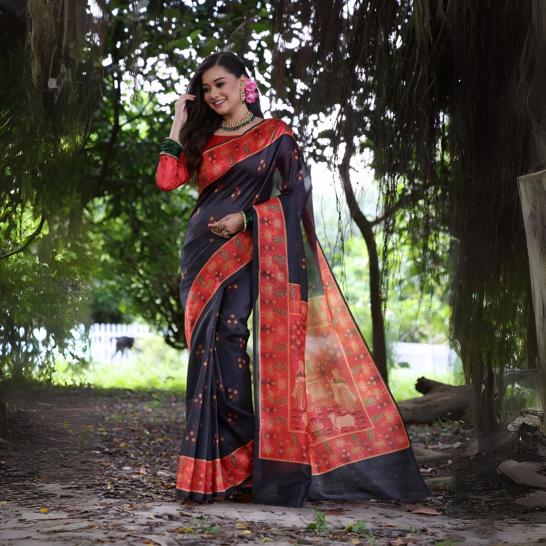 Woman wearing a black silk saree with a red border and traditional motifs, paired with a red blouse and gold jewelry, standing on a forest path surrounded by lush greenery