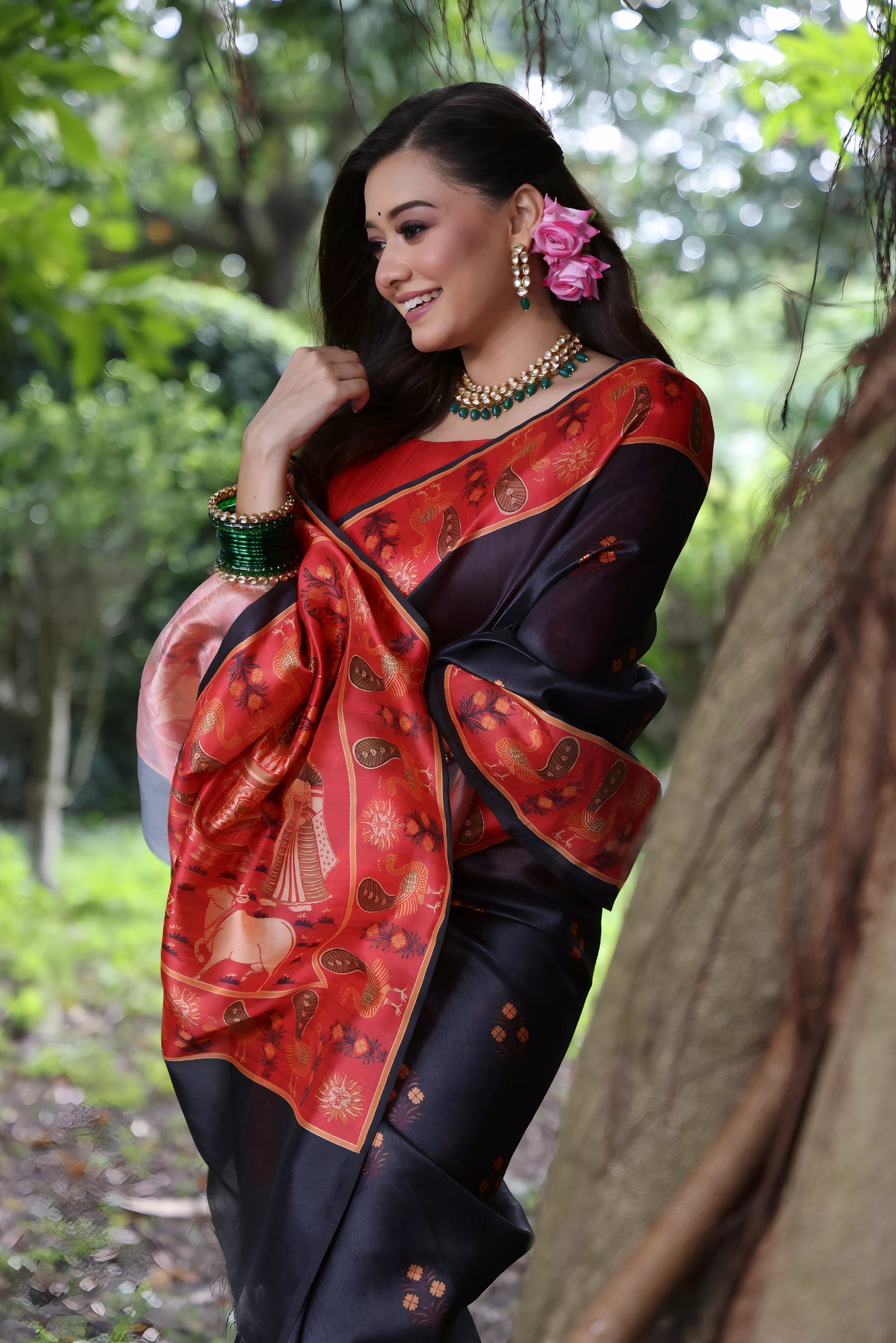 Woman wearing a black silk saree with a red border and traditional motifs, paired with a red blouse and gold jewelry, standing on a forest path surrounded by lush greenery