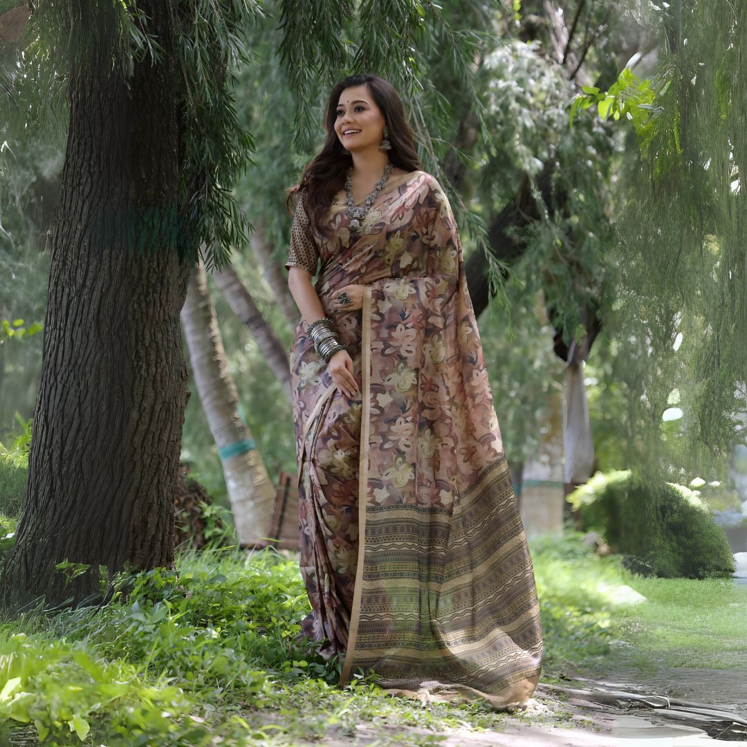 Woman wearing a brown floral silk saree with a tribal patterned border and silver jewelry, standing outdoors beside a large tree in a lush green garden.
