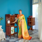 Woman in a green and orange silk saree with gold jewelry standing beside a vintage wooden cabinet in a stylish room with blue walls and marble flooring.