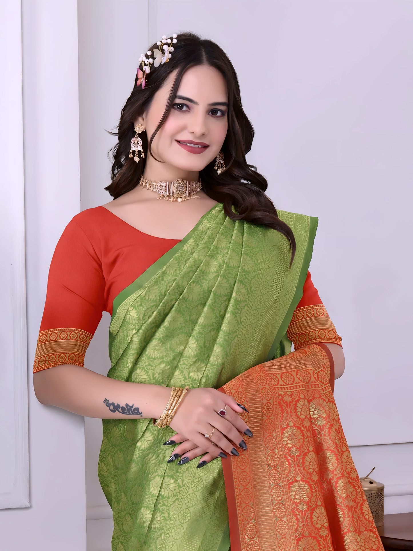 Woman wearing a green silk saree with an orange pallu and red blouse, accessorized with traditional jewelry, standing indoors near decorative plants and a wooden console table
