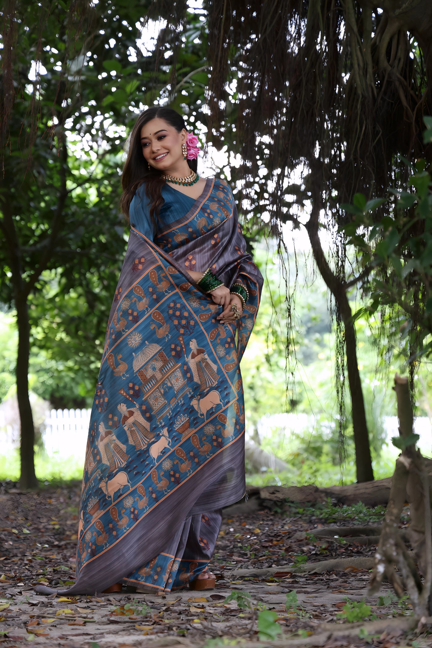 Woman wearing a grey silk saree with a blue border featuring traditional Pattachitra art and motifs, paired with green bangles and statement jewelry, standing on a leafy forest path under dense trees