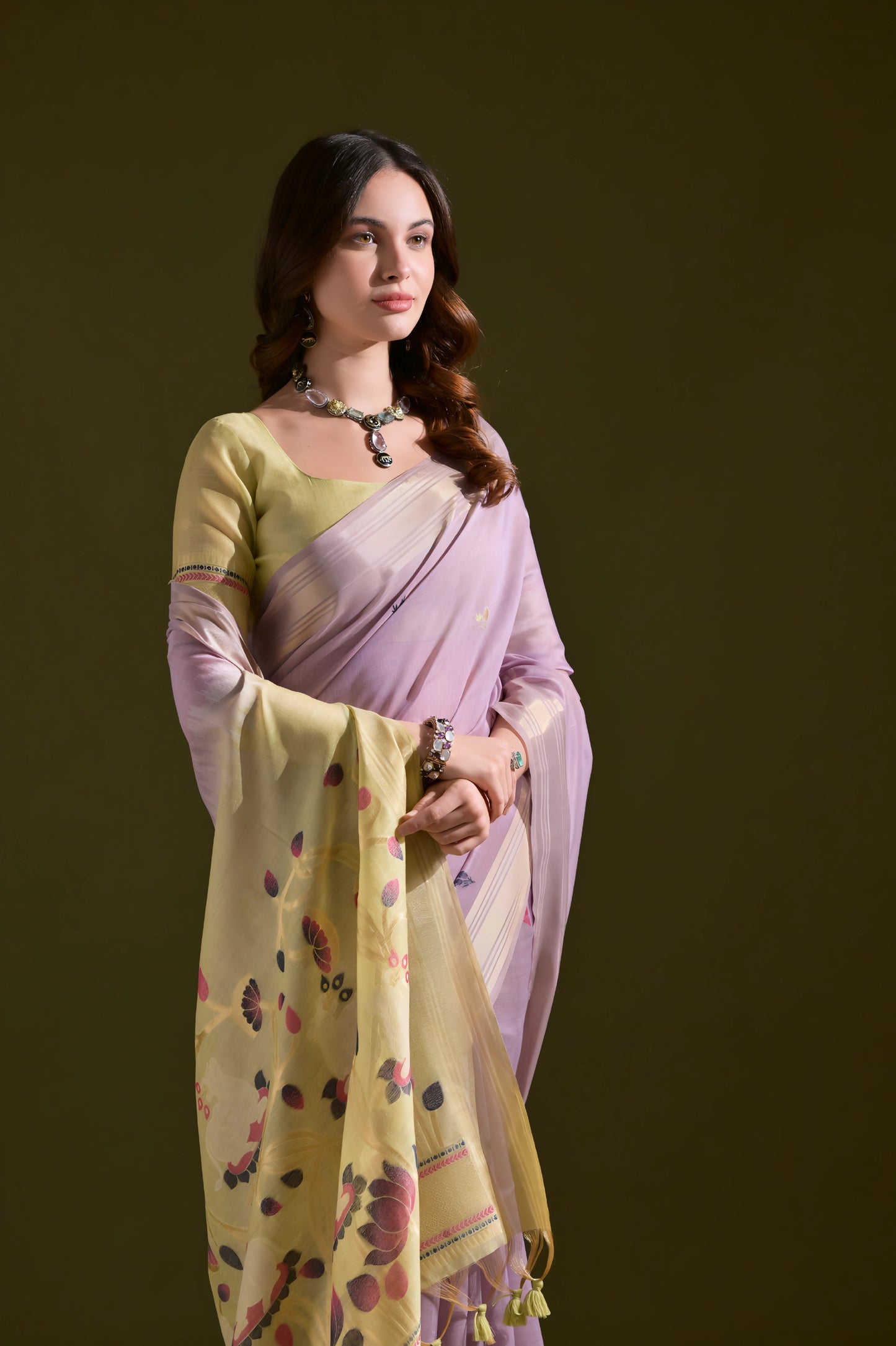 Woman wearing a lavender silk saree with a yellow floral pallu and tassel border, paired with a matching blouse and silver necklace, standing against an olive green background.
