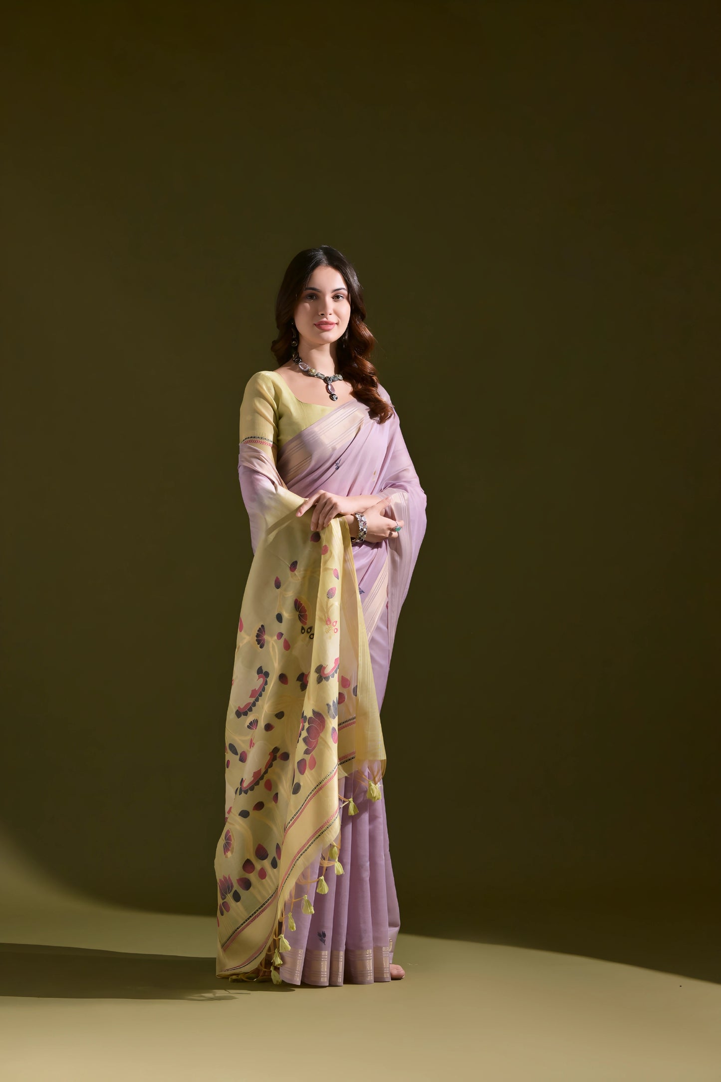 Woman wearing a lavender silk saree with a yellow floral pallu featuring tassel details, paired with a matching blouse and silver jewelry, standing against an olive green background.