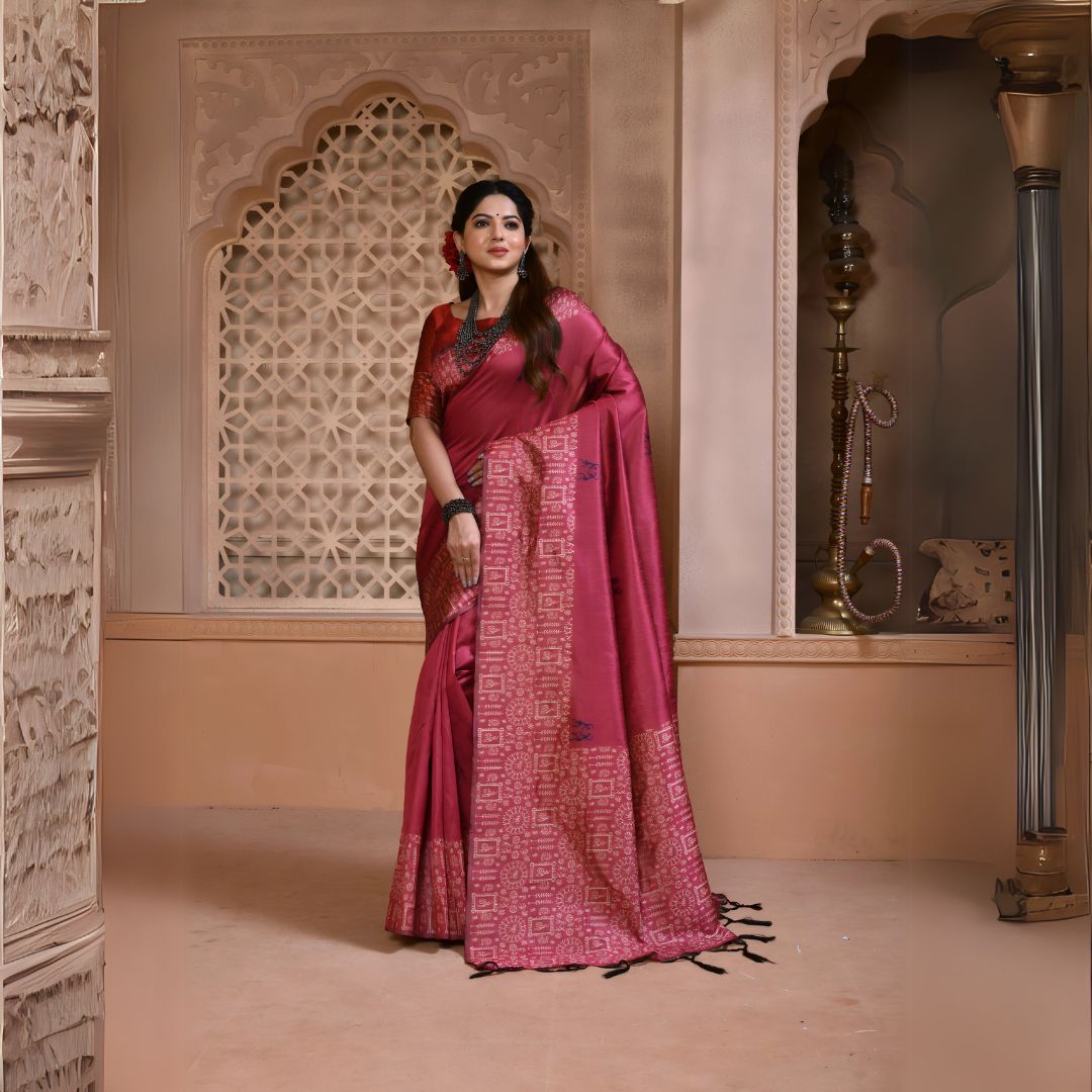 Woman wearing a maroon silk saree with a geometric patterned pallu and traditional jewelry, standing in a heritage-inspired room with ornate carved walls and decorative.
