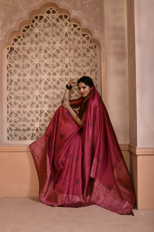 Woman wearing a maroon silk saree with a geometric patterned pallu and traditional jewelry, standing in a heritage-inspired room with ornate carved walls and decorative window lattice