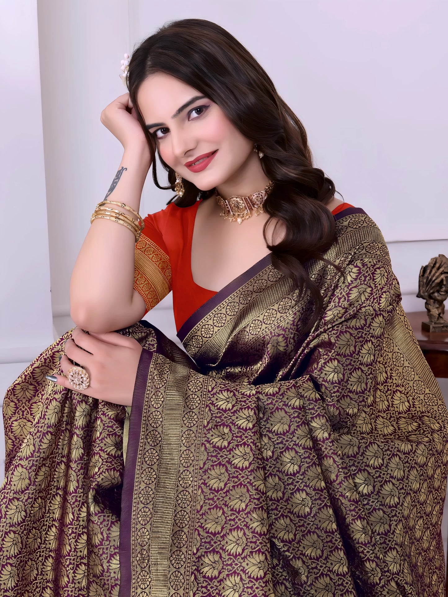 Woman wearing an olive green silk saree with intricate gold motifs and an orange border, paired with a red blouse and traditional jewelry, standing indoors beside a potted plant and wooden console table