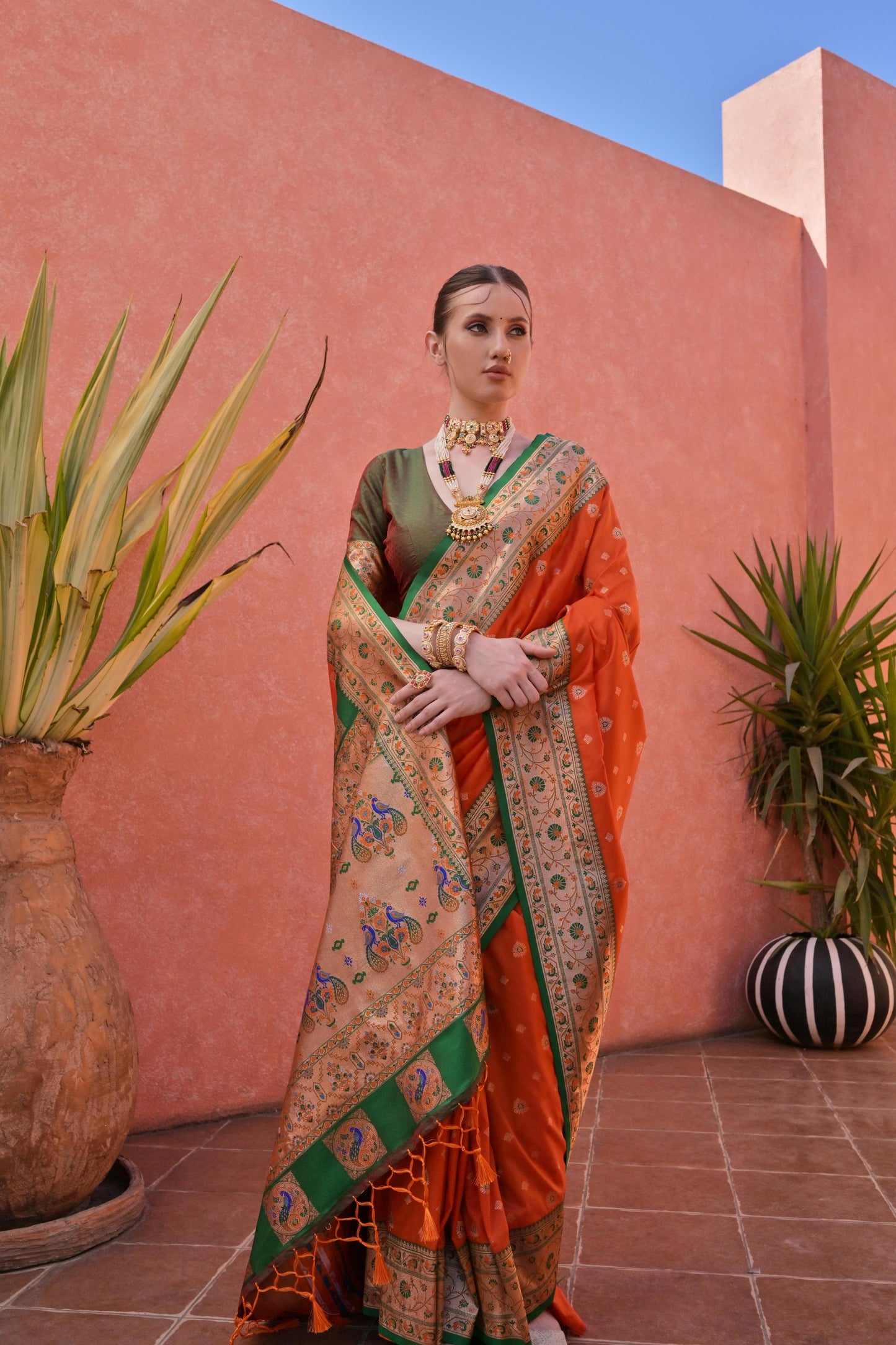 Woman wearing an orange and green silk saree with intricate peacock border and traditional gold jewelry, standing in front of a terracotta wall with potted plants.