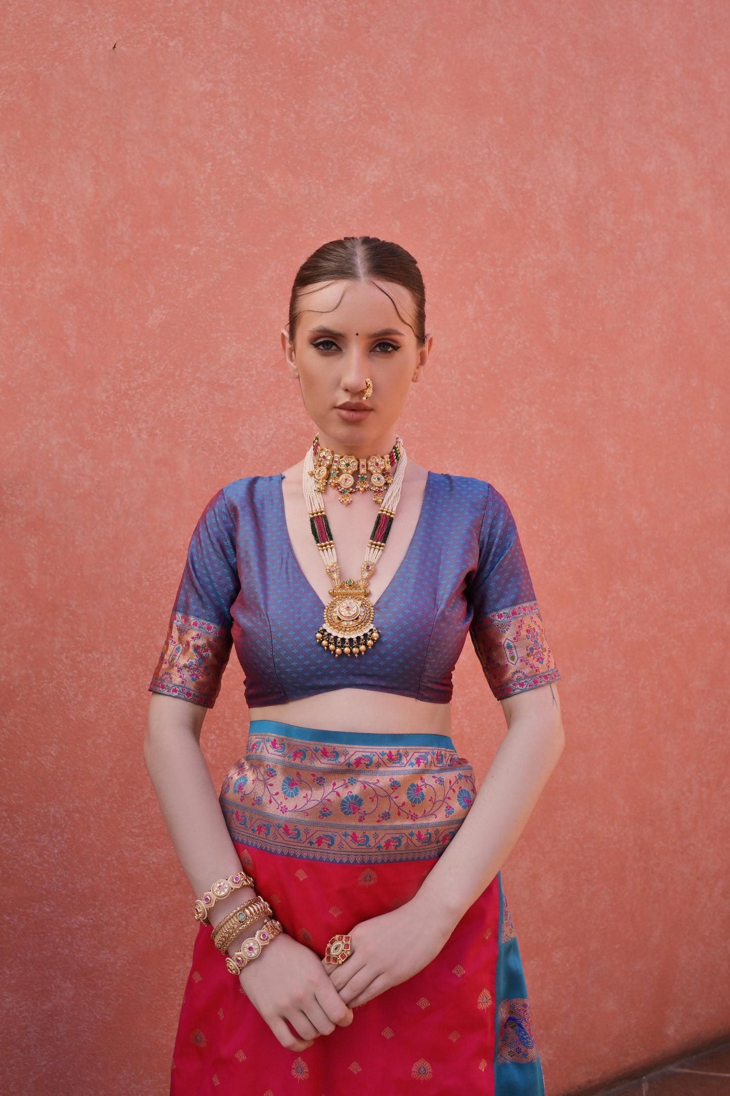 Woman wearing a pink and blue silk saree with intricate patterns and gold jewelry, standing in front of a terracotta wall with potted green plants under a clear blue sky.