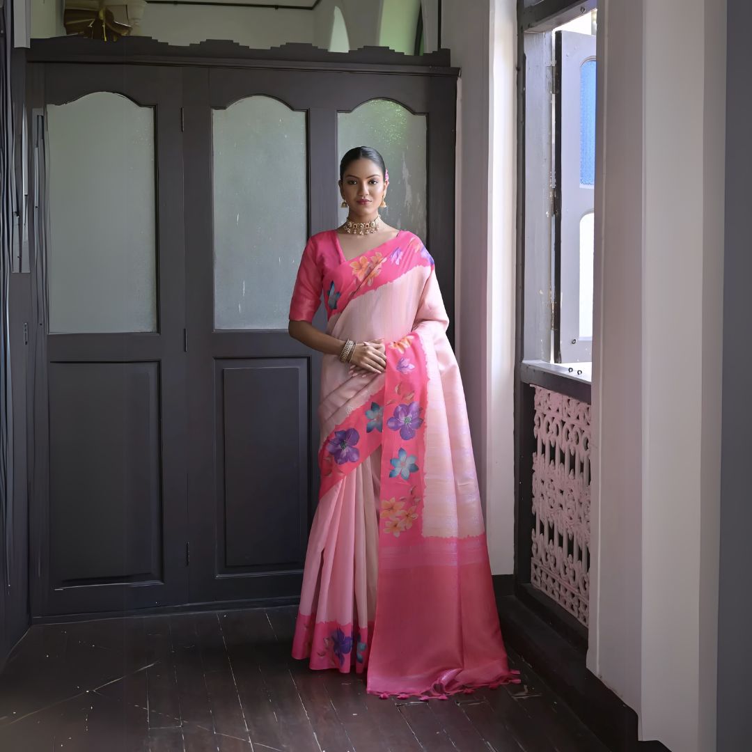 Woman wearing a pink silk saree with multicolor floral patterns and traditional jewelry, standing indoors near a wooden door and window with natural light.