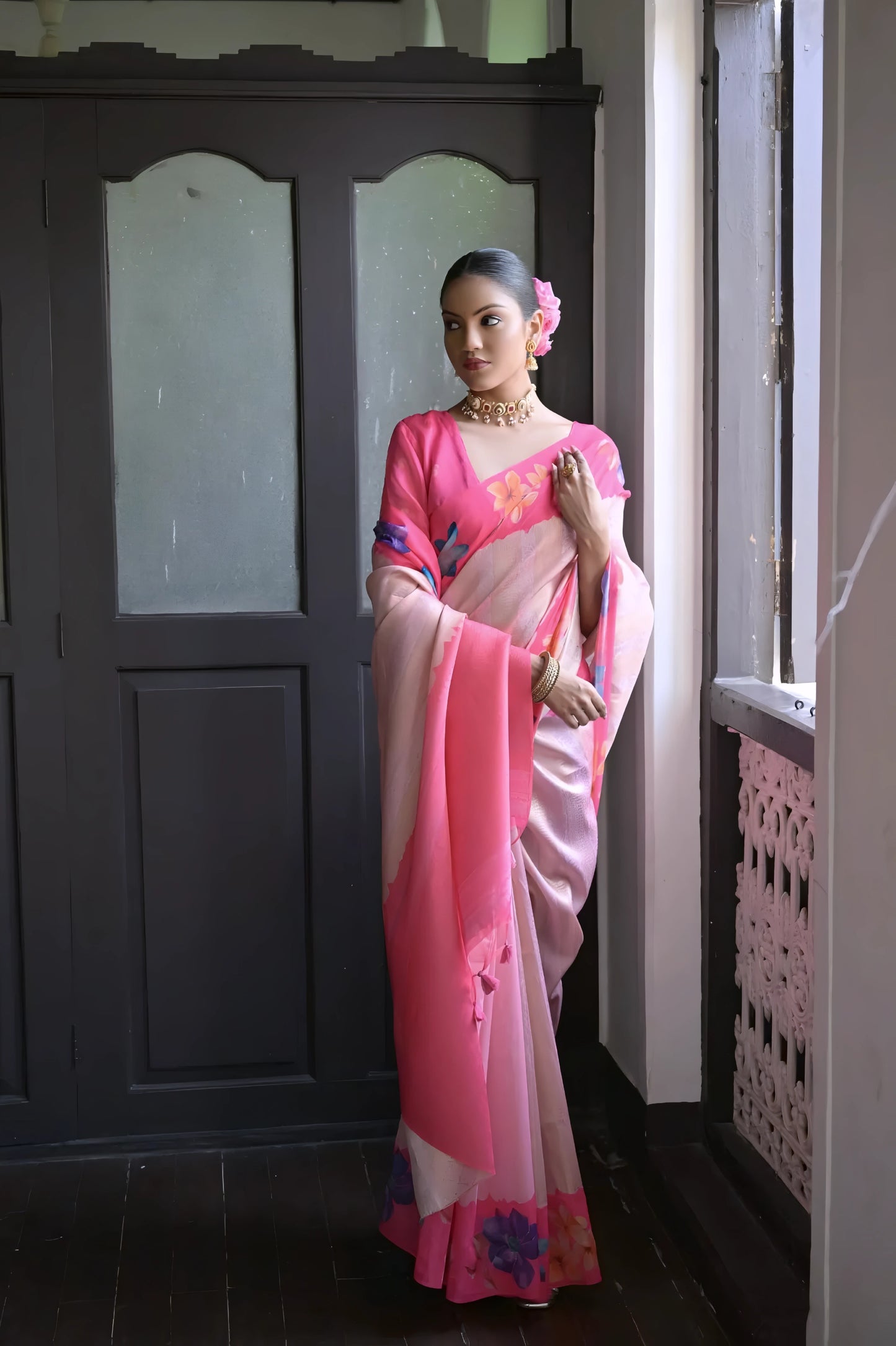 Woman wearing a pink silk saree with multicolor floral patterns and traditional jewelry, standing indoors near a wooden door and window with natural light