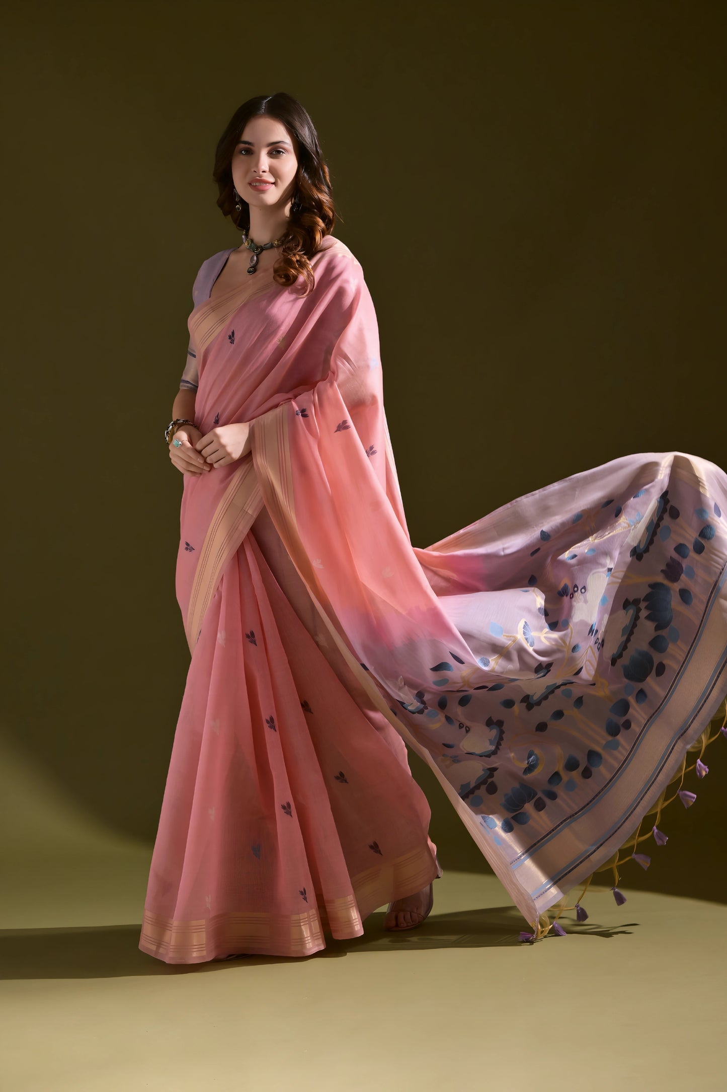 Woman wearing a pink silk saree with a blue floral pallu and tassel border, paired with a matching blouse and silver necklace, standing against an olive green background.