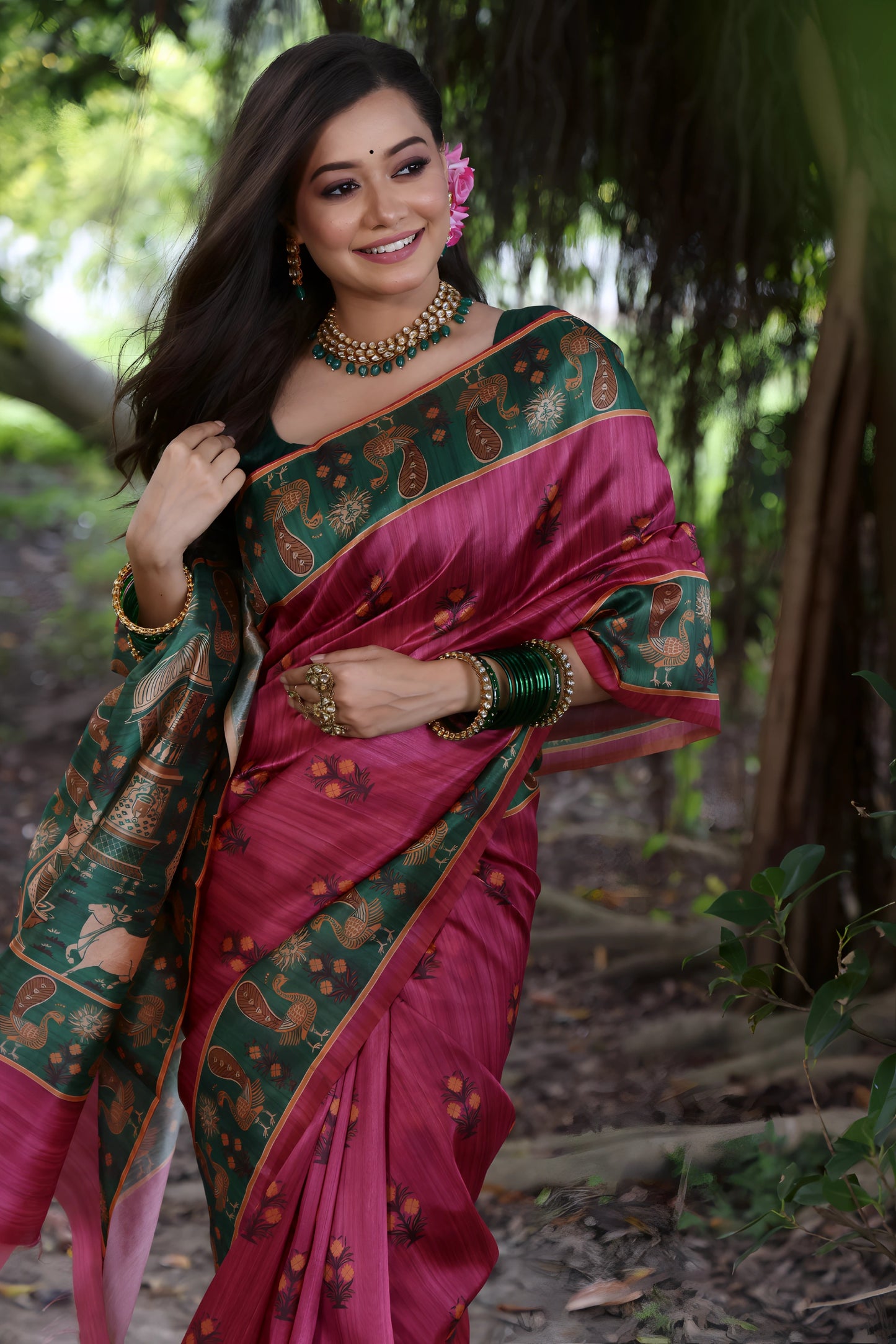 Woman wearing a pink silk saree with a green pallu featuring traditional motifs and gold jewelry, standing on a forest path surrounded by lush greenery