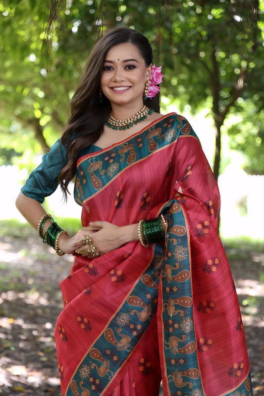 Woman wearing a pink silk saree with a teal blue border and traditional motifs, paired with a teal blouse, green bangles, and floral hair accessory, standing on a forest path under lush green trees