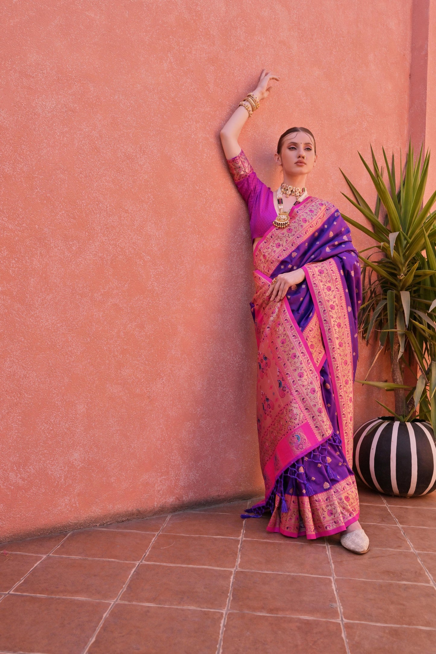 Woman in a purple and pink silk saree with gold jewelry posing by a terracotta wall next to a potted green plant.
