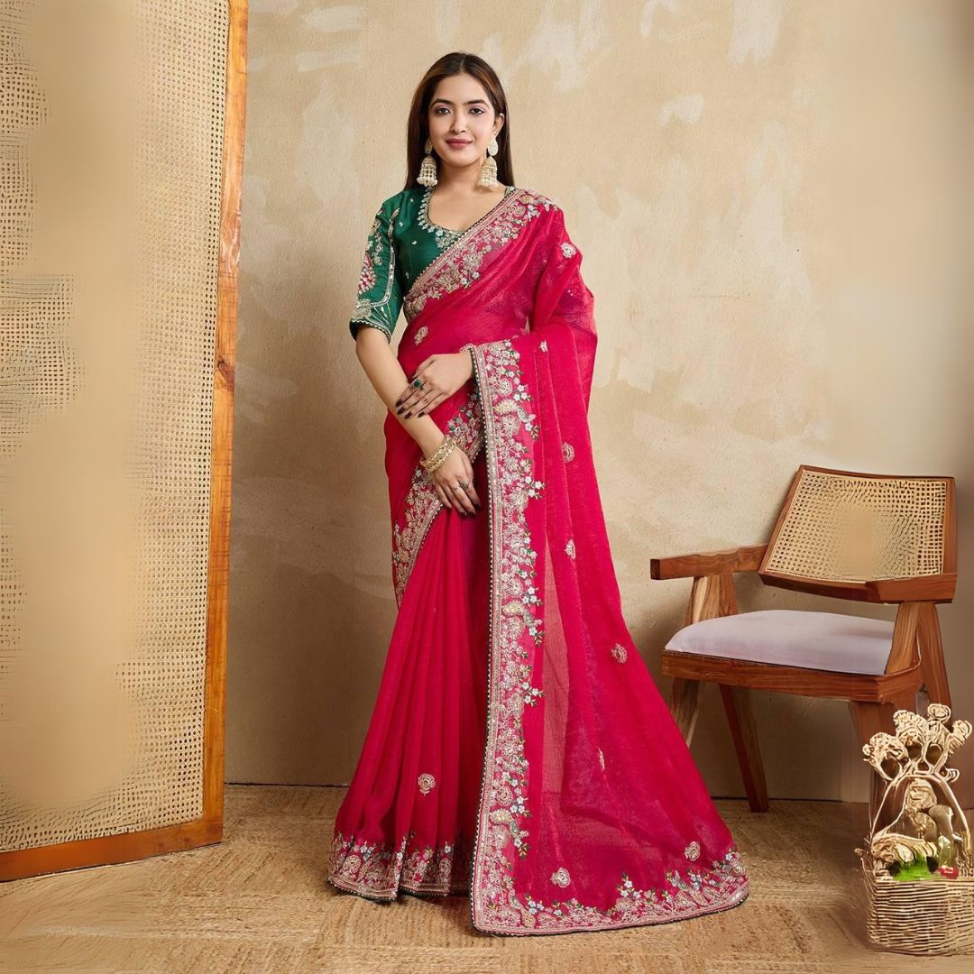 Woman wearing a royal purple silk saree with intricate pink and gold border and pallu, paired with a matching blouse and traditional gold jewelry, standing on a terracotta-tiled outdoor terrace with decorative plants and peach-colored walls