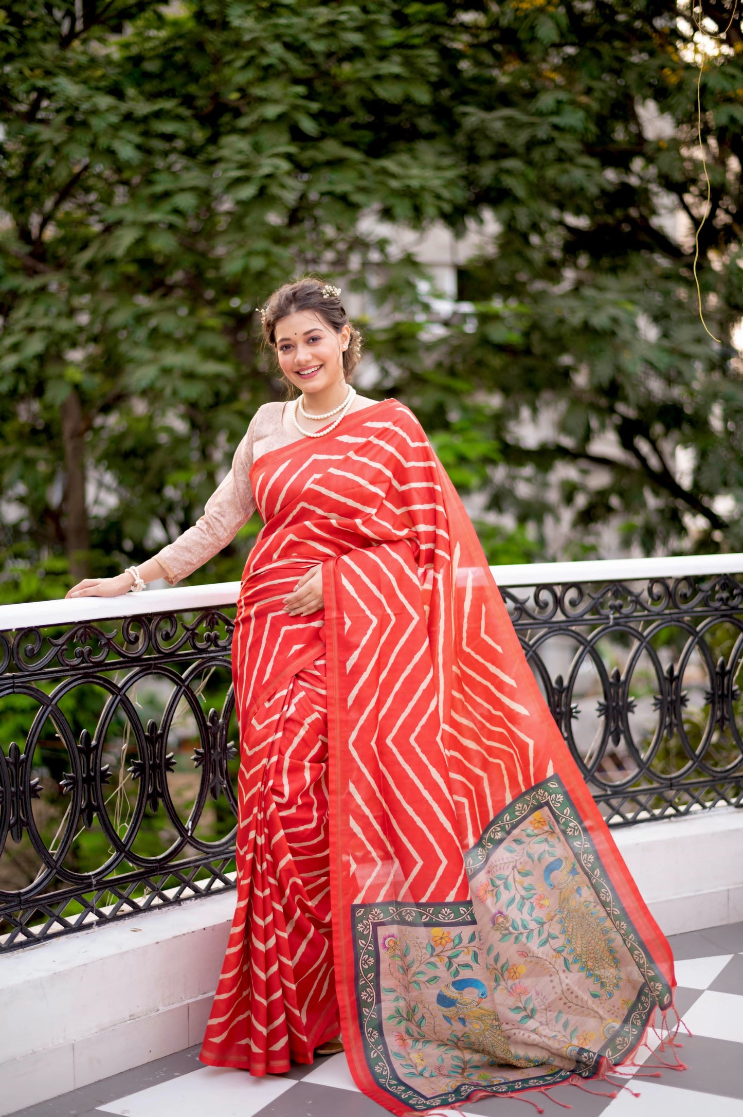 Woman wearing a red saree with bold white geometric stripes and a floral embroidered pallu, standing on a balcony with decorative railing and lush green garden in the background