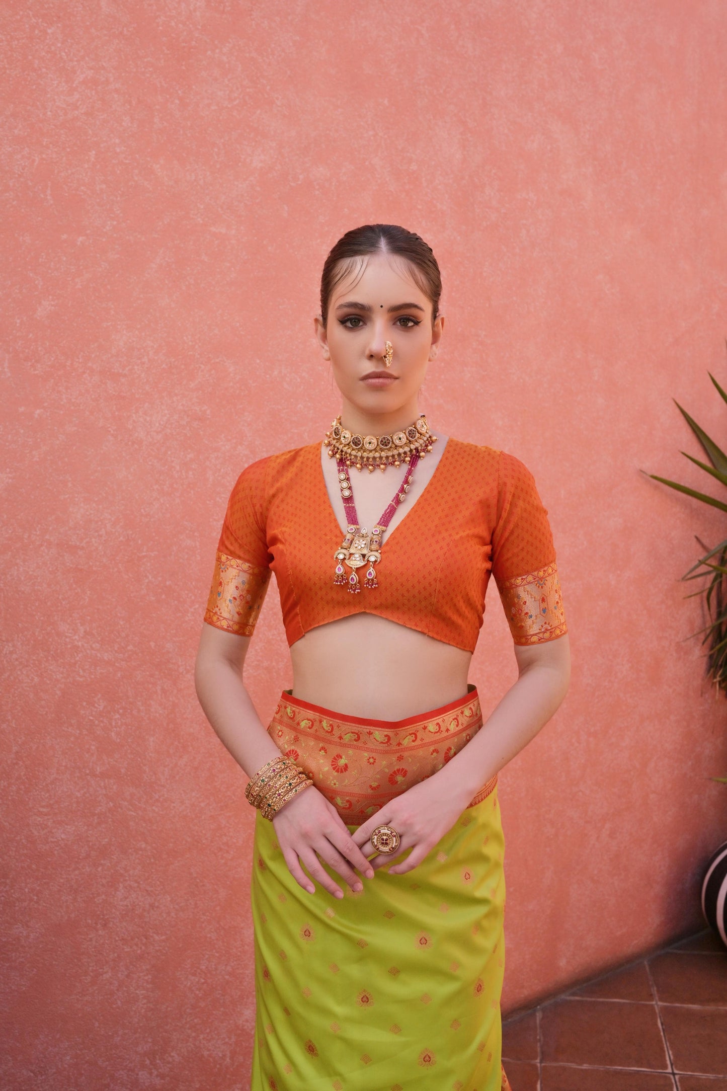 Woman wearing a green and orange traditional saree with matching blouse and jewelry, standing in a stylish courtyard with potted plants and pink walls