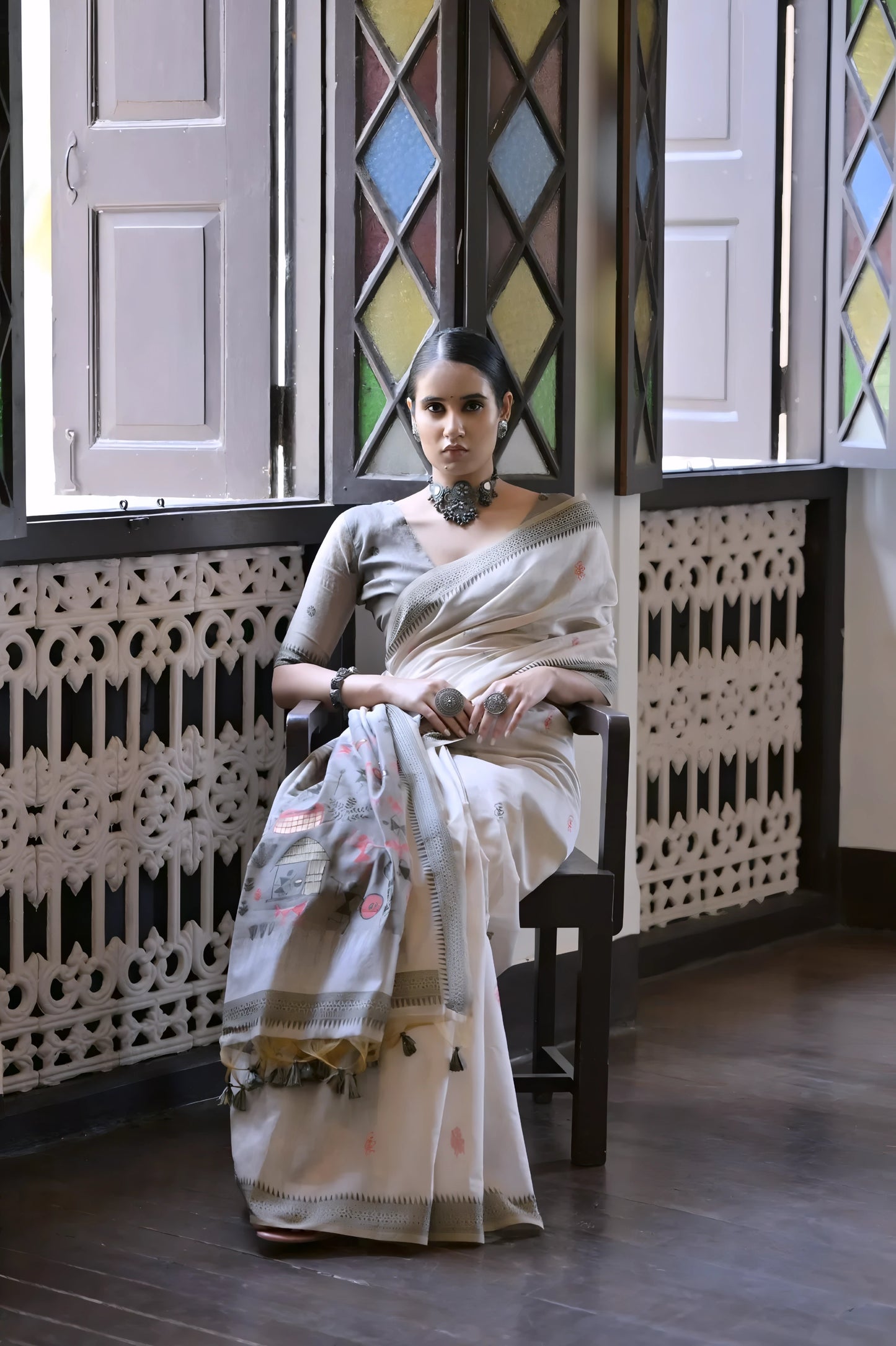 Woman wearing a white silk saree with handpainted motifs and a tassel border, accessorized with traditional jewelry, standing indoors beside vintage stained glass windows with pastel light