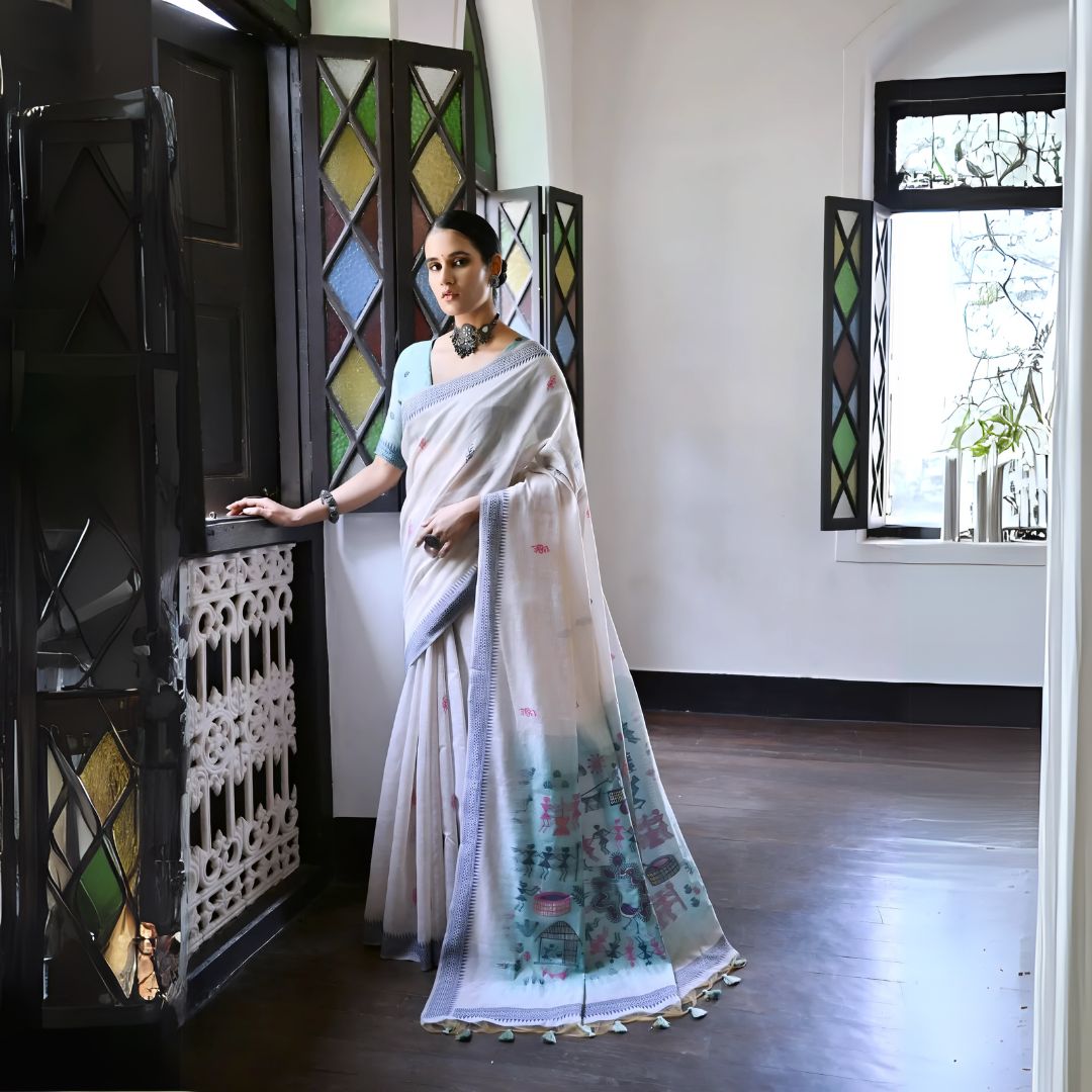 Woman wearing a white silk saree with pastel village motifs and a tassel border, paired with a light blue blouse and statement necklace, standing indoors beside vintage stained glass windows.