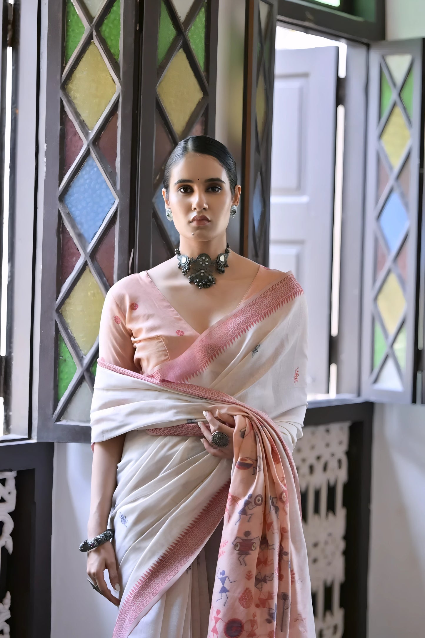Woman wearing a white silk saree with a pink blouse and floral pallu, accessorized with a statement necklace, standing indoors beside vintage stained glass windows