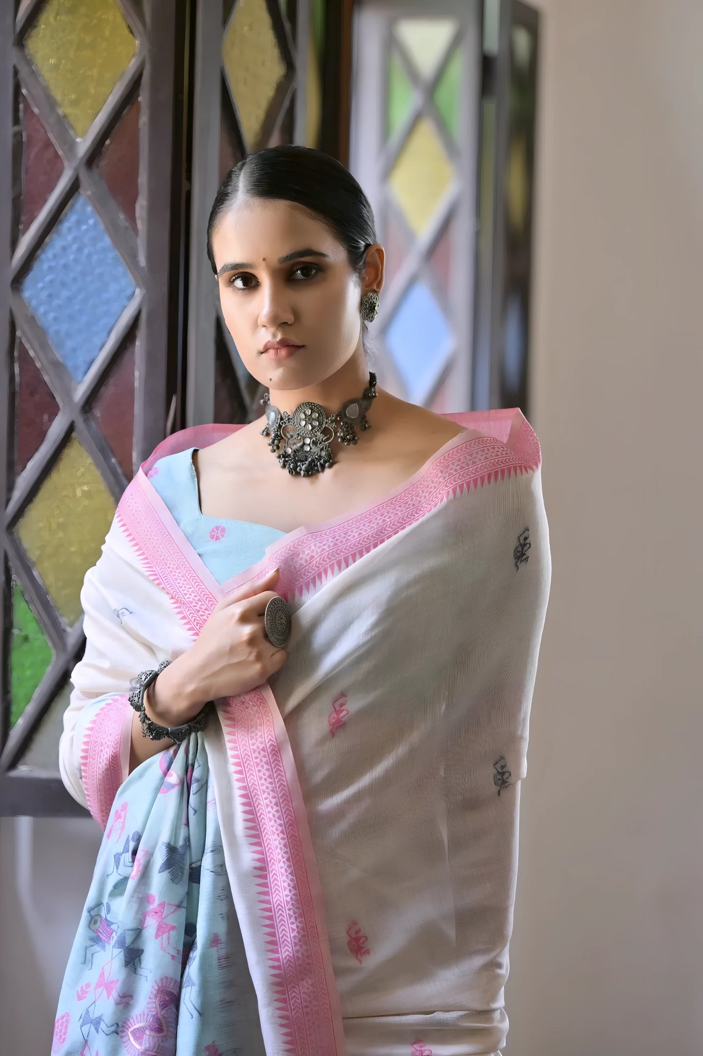 Woman wearing a white silk saree with pastel pink and blue village motifs and a tassel border, paired with a pink and blue blouse and statement necklace, standing indoors beside vintage stained glass windows