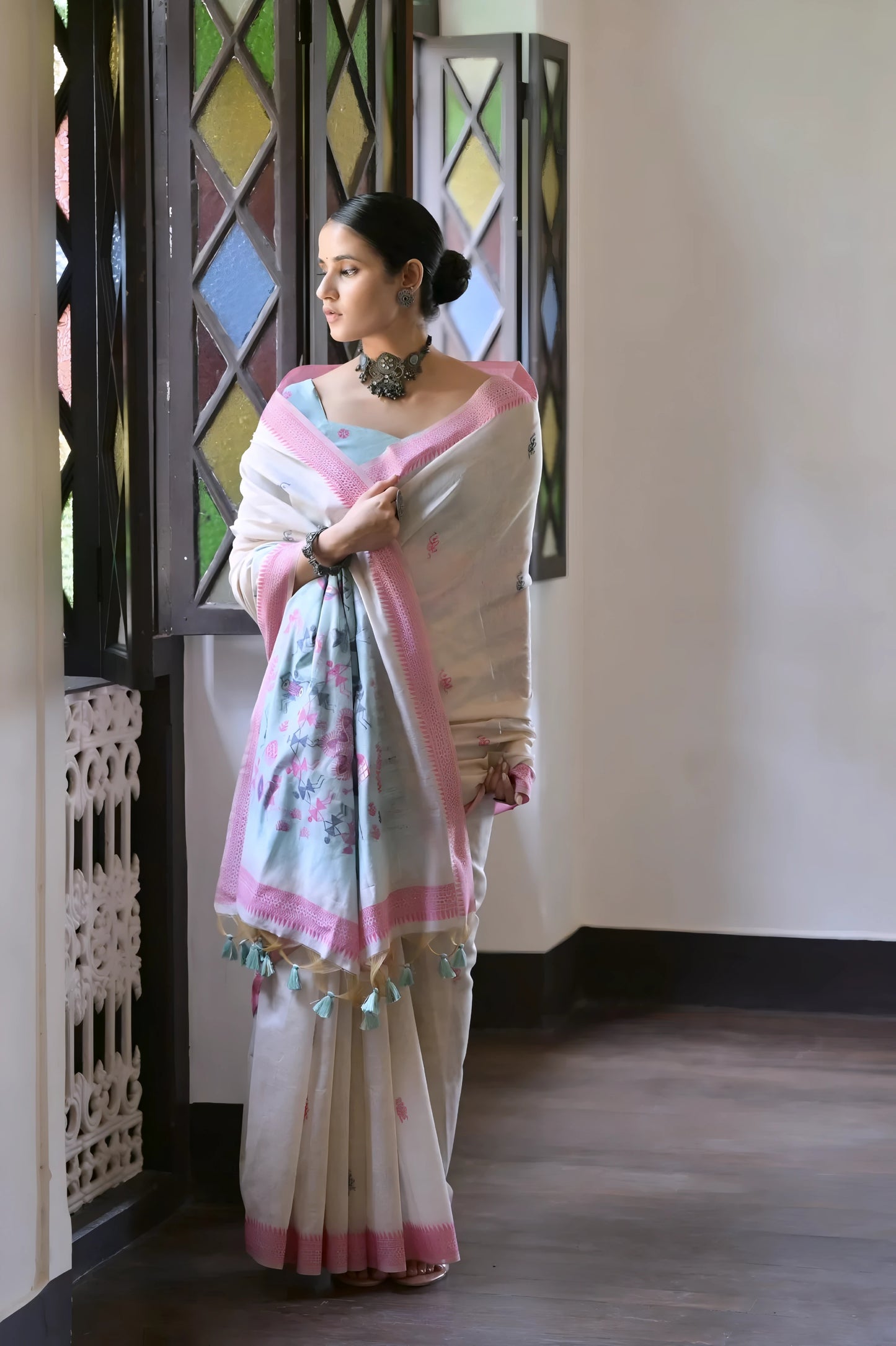 Woman wearing a white silk saree with pastel pink and blue village motifs and a tassel border, paired with a pink and blue blouse and statement necklace, standing indoors beside vintage stained glass windows