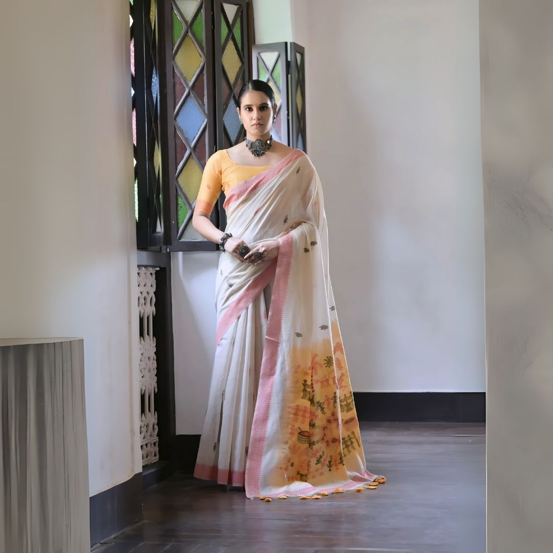 Woman wearing a white silk saree with pastel village motifs and a tassel border, paired with a yellow blouse and statement jewelry, standing indoors beside vintage stained glass windows