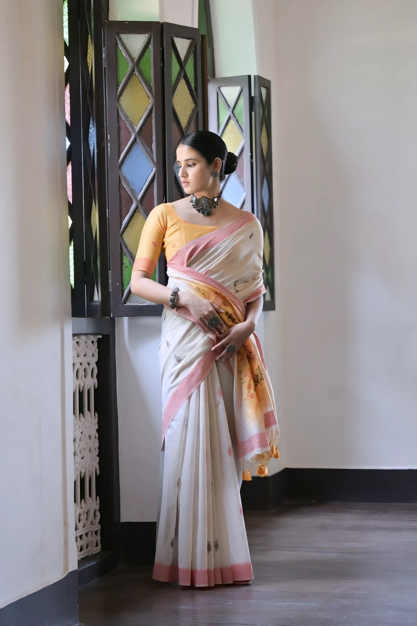 Woman wearing a white silk saree with pastel village motifs and a tassel border, paired with a yellow blouse and statement jewelry, standing indoors beside vintage stained glass windows.