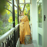 Woman wearing a yellow saree with bold white geometric stripes and a floral embroidered pallu, accessorized with pearl jewelry, standing on a balcony with decorative railing overlooking a lush green garden