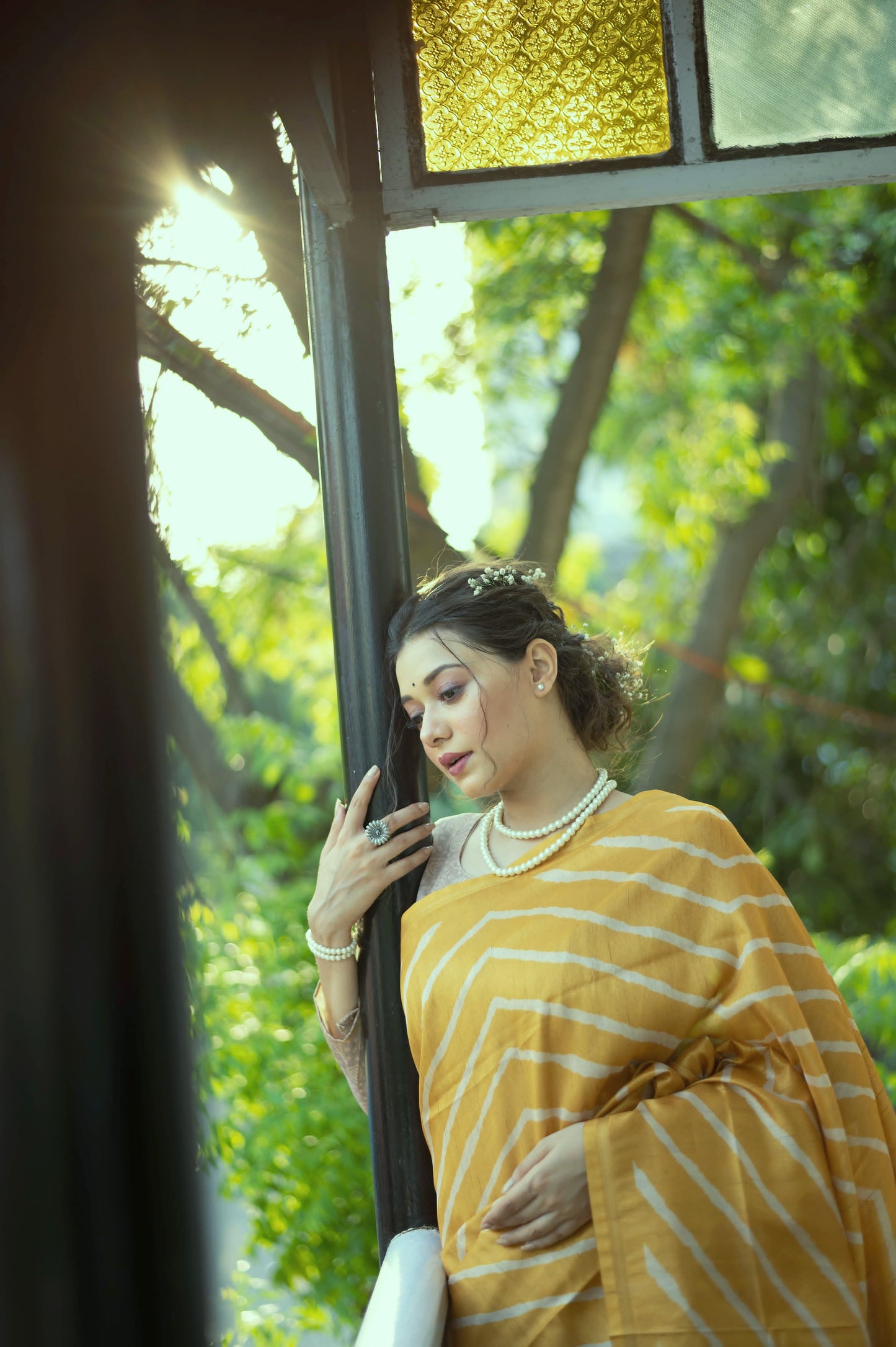 Woman wearing a yellow saree with bold white geometric stripes and a floral embroidered pallu, accessorized with pearl jewelry, standing on a balcony with decorative railing overlooking a lush green garden.