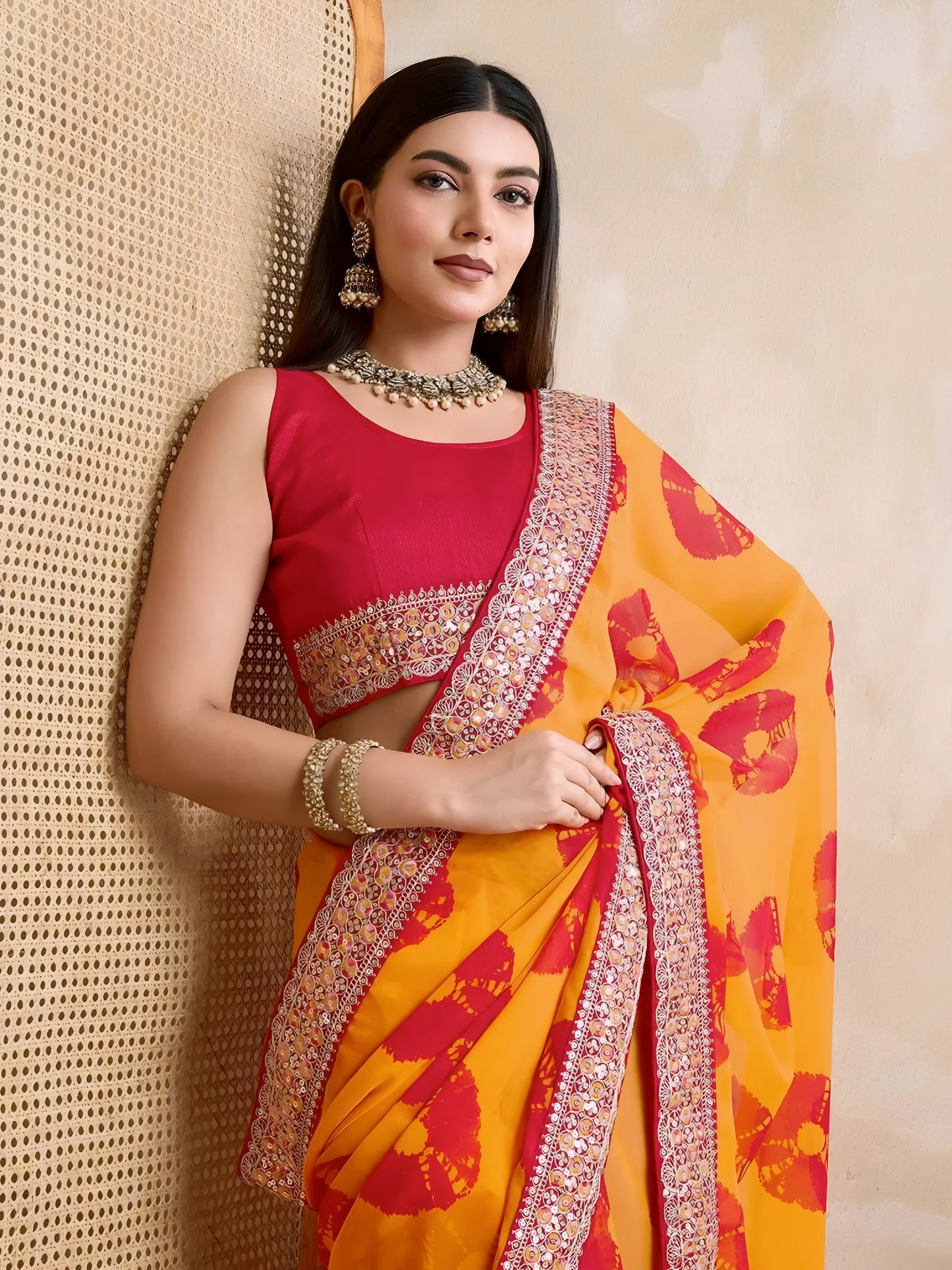 Woman wearing a yellow georgette saree with red floral patterns and an embellished pink border, paired with a sleeveless pink blouse and traditional jewelry, standing beside a wooden cane screen and modern chair against a beige wall.