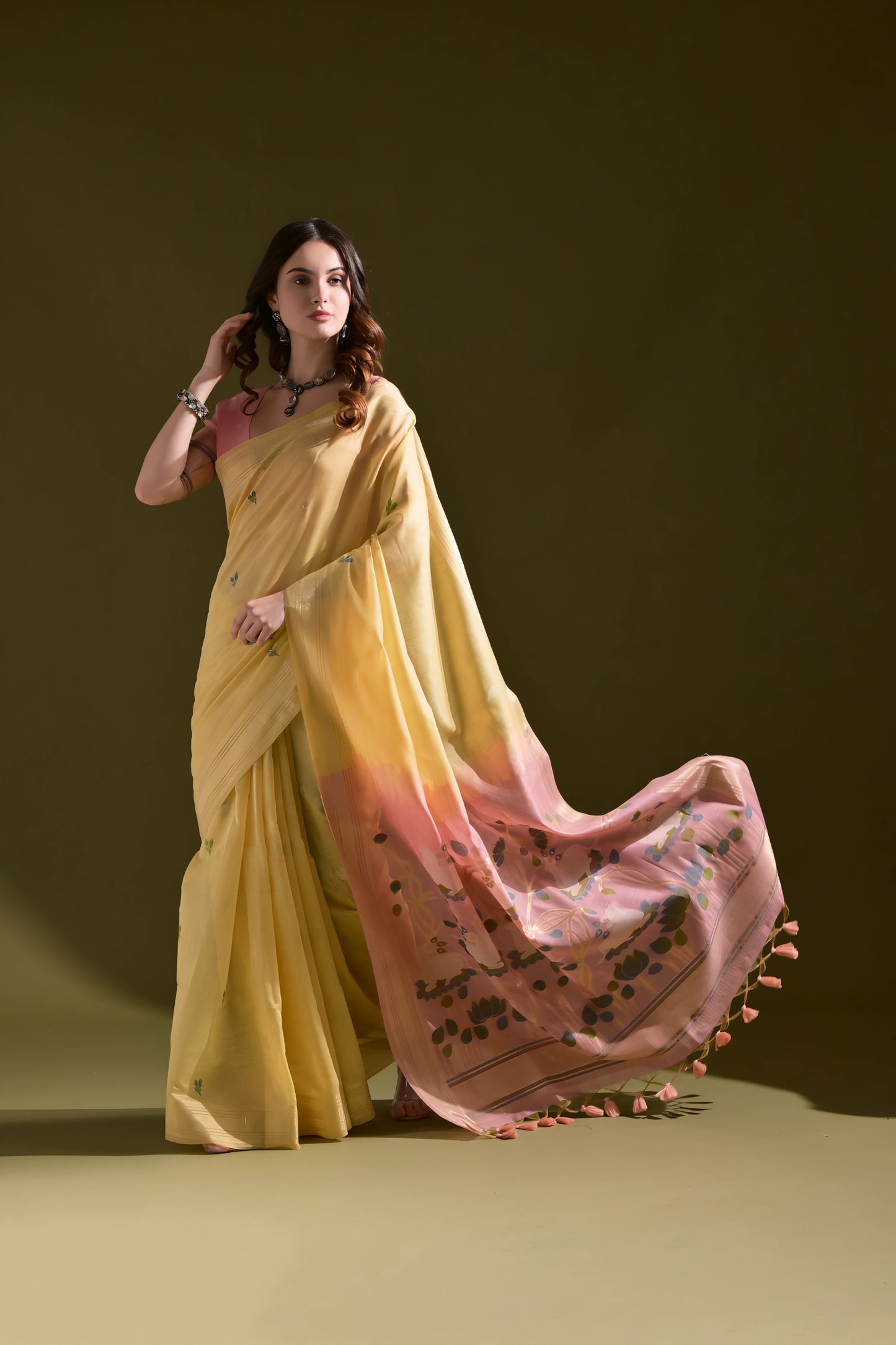 Woman wearing a yellow silk saree with a pink floral pallu and tassel border, paired with a pink blouse and silver jewelry, standing against an olive green background.