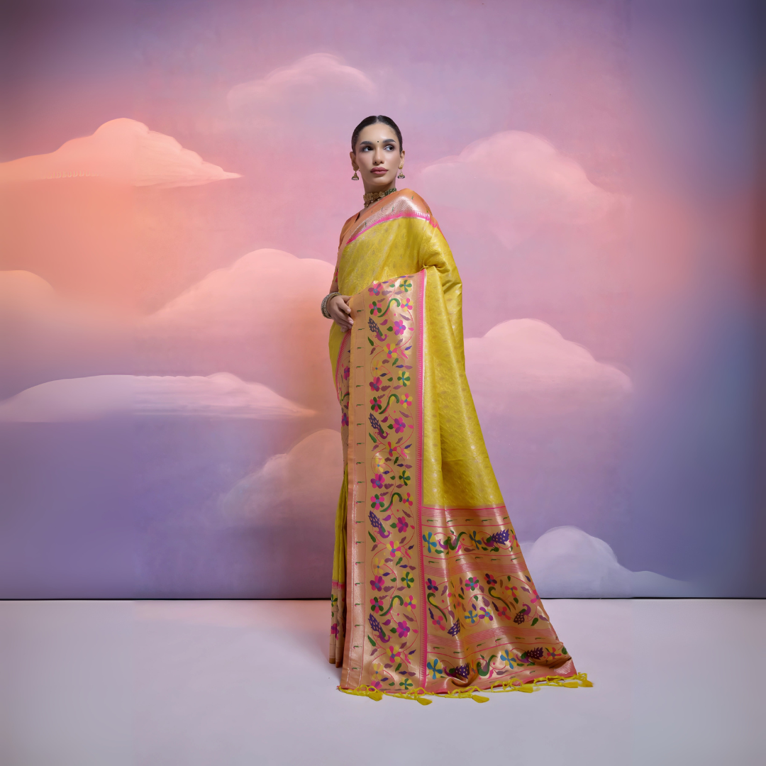 Woman wearing a yellow Banarasi silk saree with intricate pink and multicolor floral zari border and pallu, paired with traditional jewelry, posing in a pastel-themed studio with a dreamy cloud background