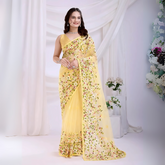 Woman wearing a yellow net saree with intricate multicolor floral embroidery and a matching sleeveless blouse, posing in a modern white studio with draped curtains and floral arrangements.